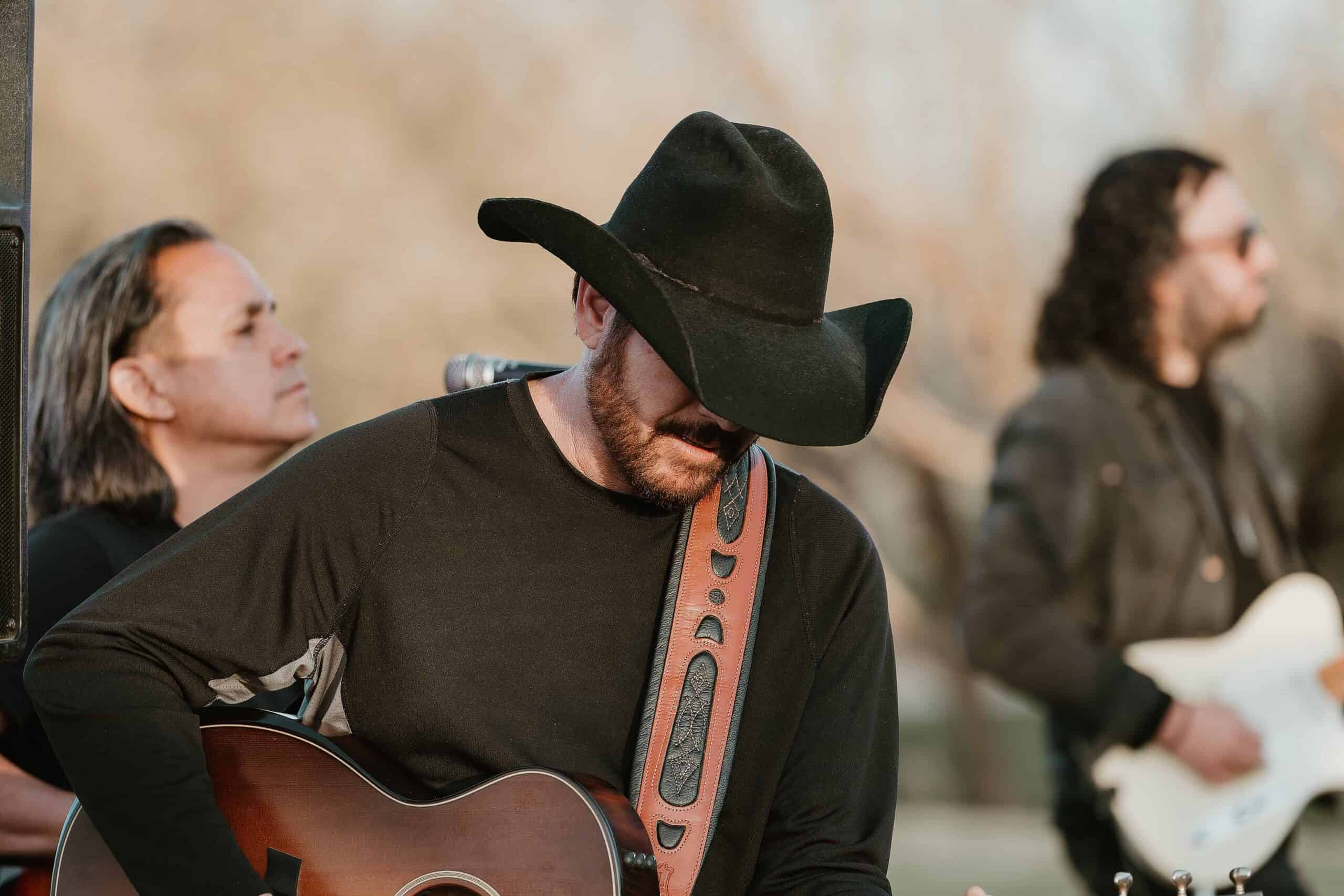 Country musician wearing a black cowboy hat performing acoustic guitar at an outdoor event in Virginia Beach.