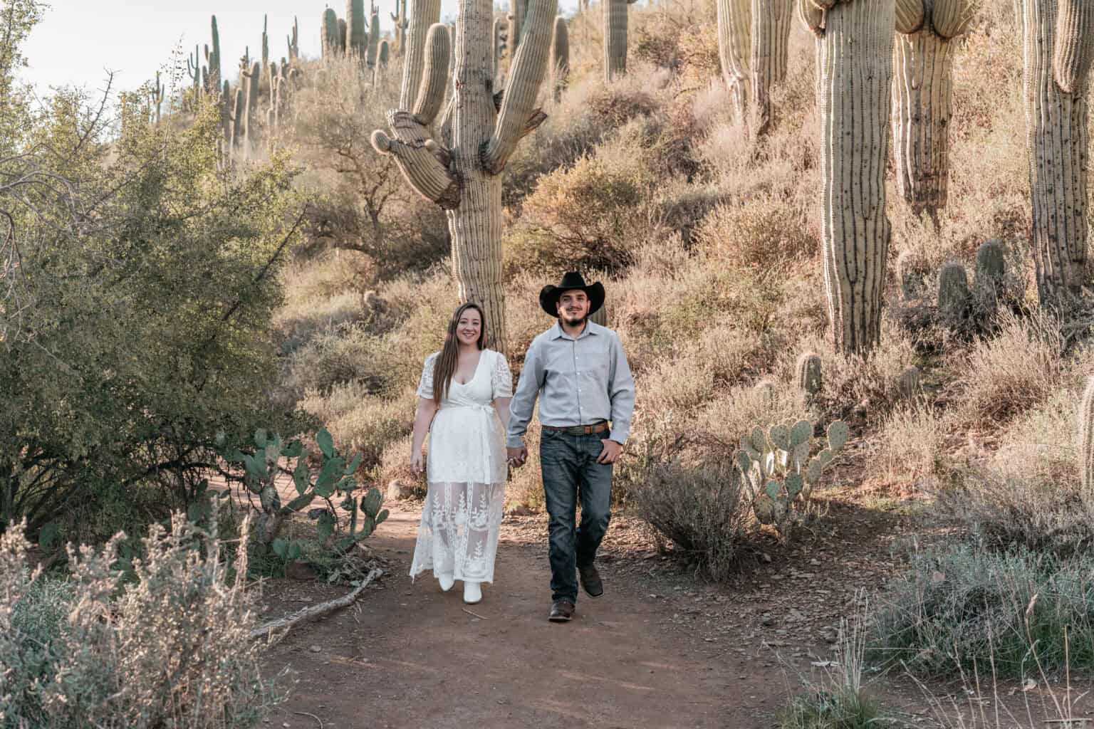 Bride in white lace dress and groom with cowboy hat walking through cactus-filled desert landscape
