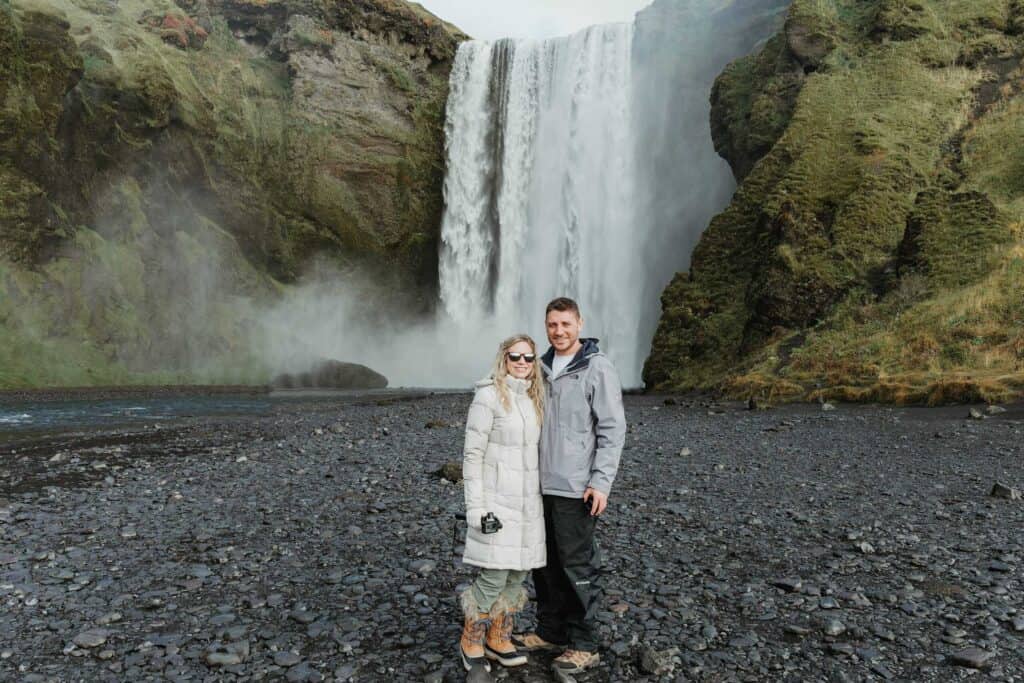 Couple posing in front of the majestic Skogafoss waterfall in Iceland, with black volcanic sand and green cliffs.