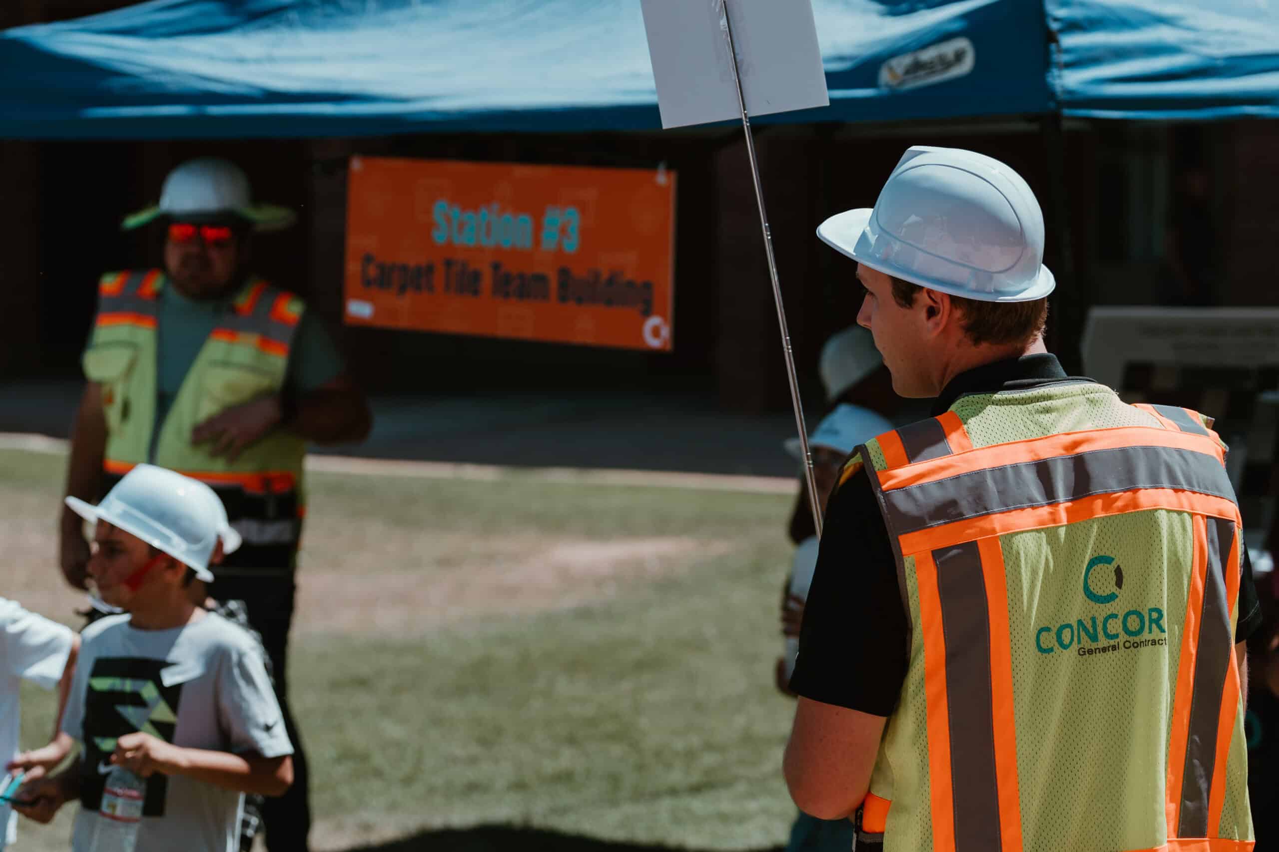 A person wearing a high-visibility vest with "CONCORD General Contractors" and a hard hat, seen from behind, participates in a "Carpet Tile Team Building" activity at a Virginia Beach event.
