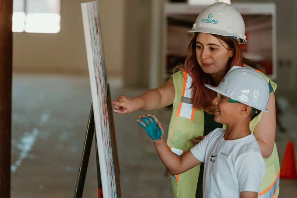 A woman in a hard hat and high-visibility vest interacts with a young boy in a hard hat, pointing at a display board, at a construction-themed event in Virginia Beach.  Taken by a Virginia Beach Videographer.
