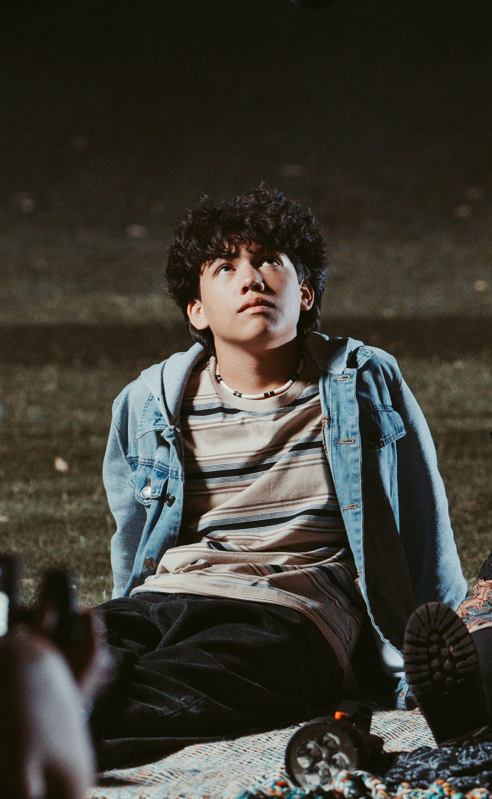 Taken by a Virginia Wedding Videographer. Young man with curly hair and a denim jacket looking up, sitting on a patterned mat in a low-light setting.