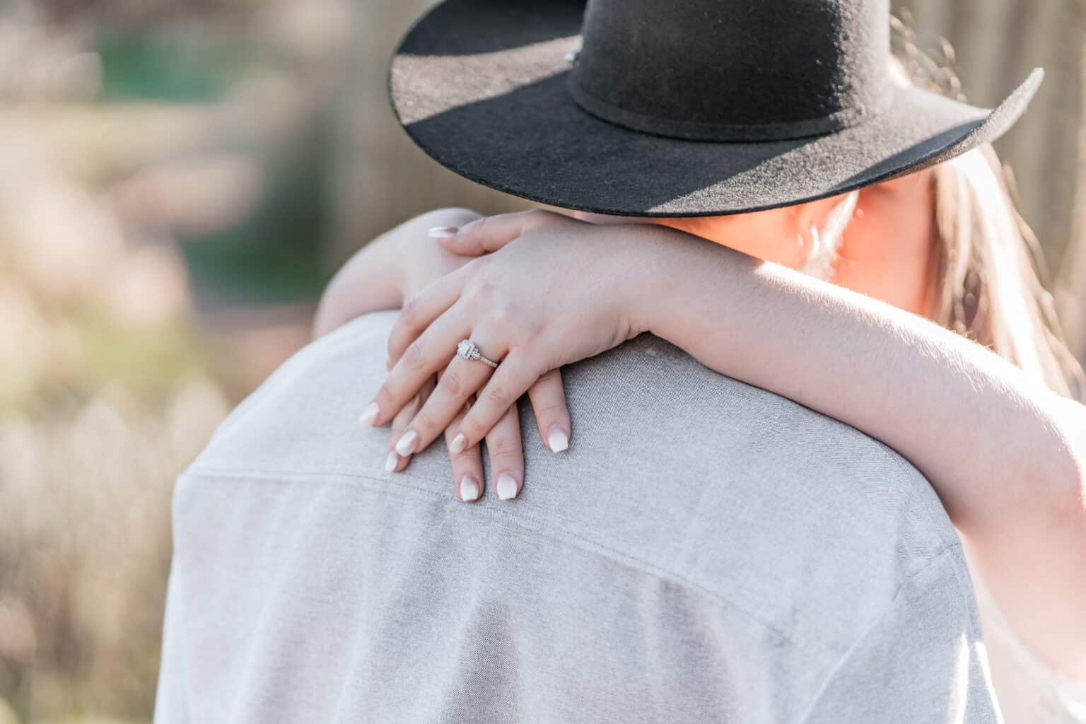 Close-up of bride's engagement ring as she embraces her fiancé during a sunlit Virginia Beach engagement session.