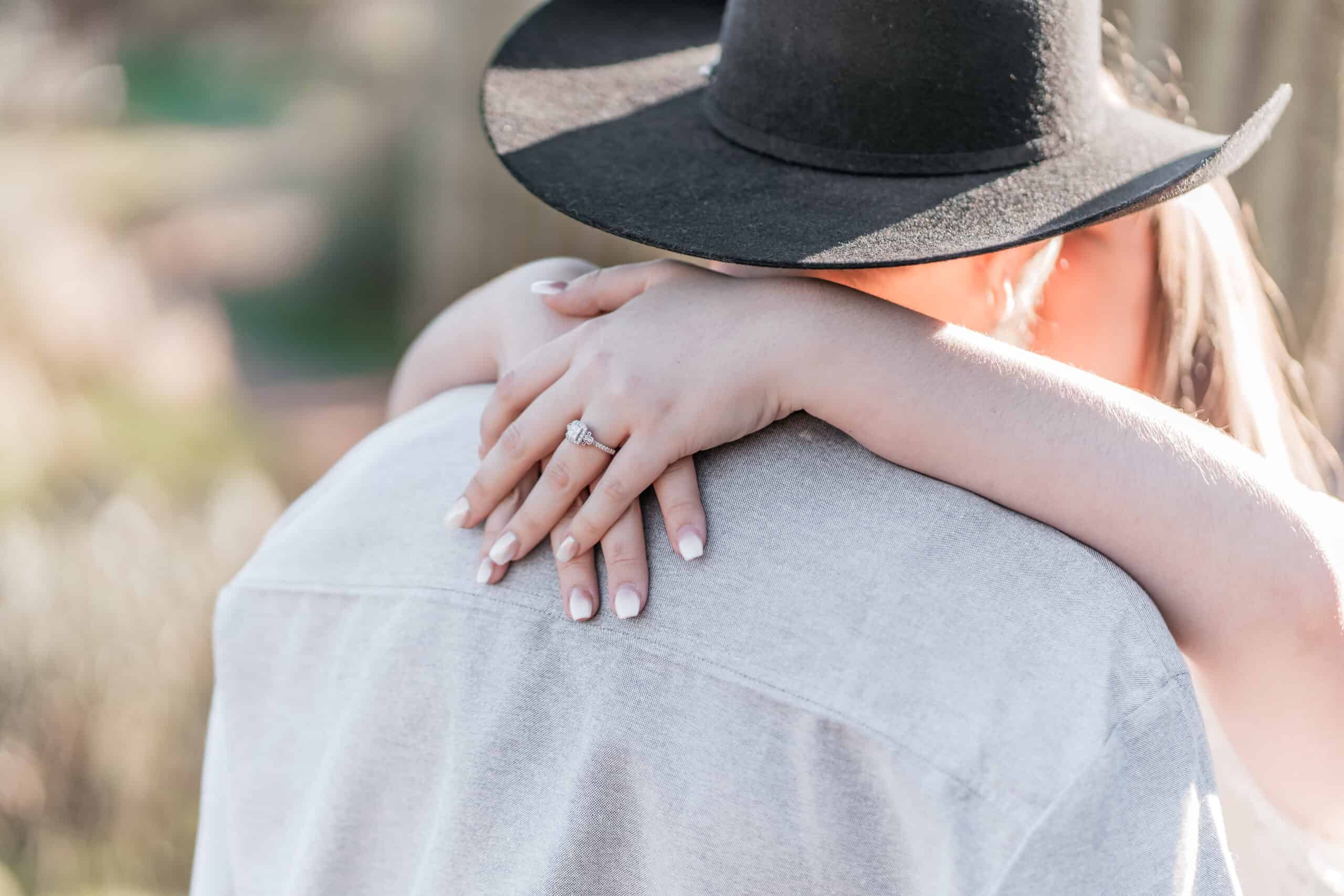Close-up of bride's engagement ring as she embraces her fiancé during a sunlit Virginia Beach engagement session.