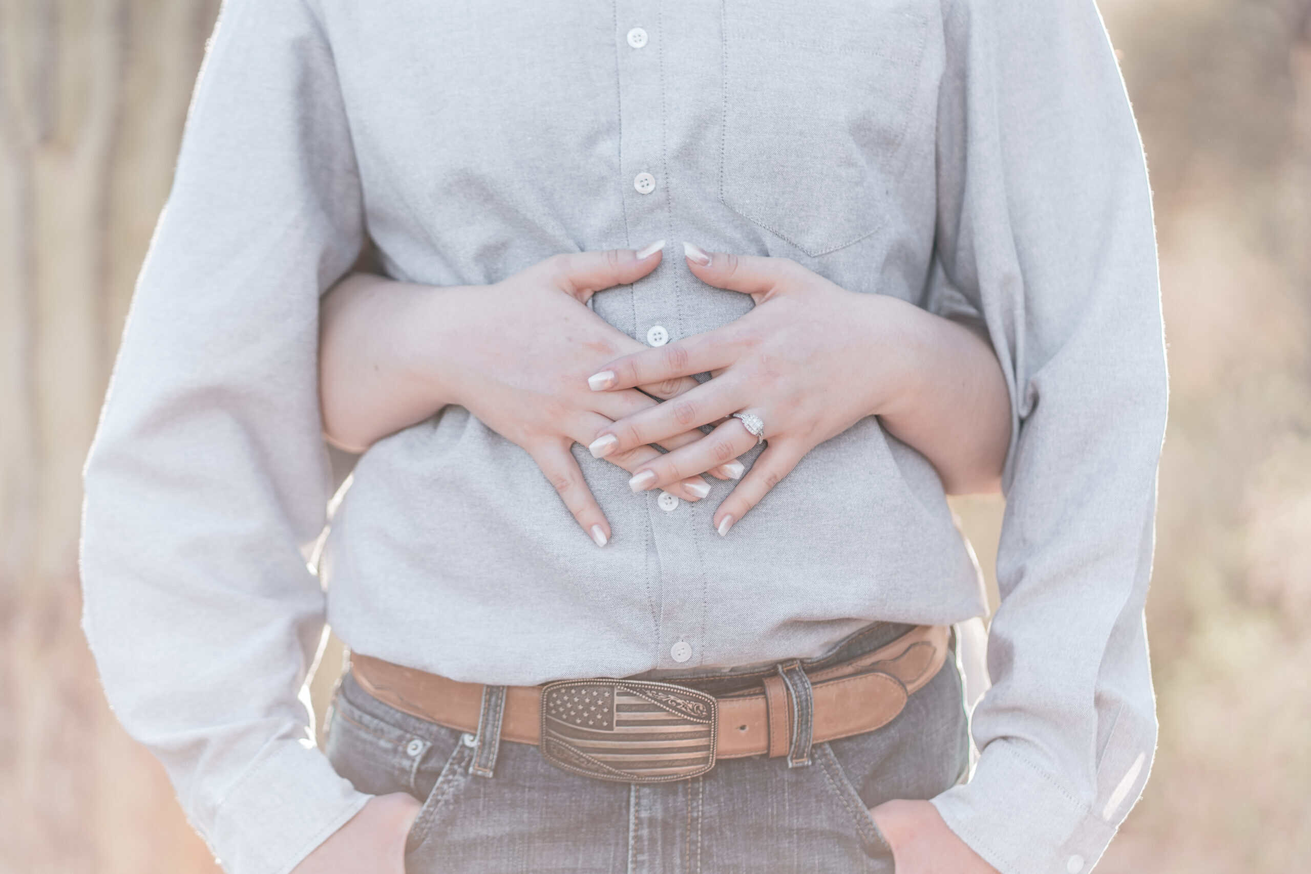 Romantic photo of bride holding fiancé from behind, showcasing her diamond engagement ring and manicured hands.