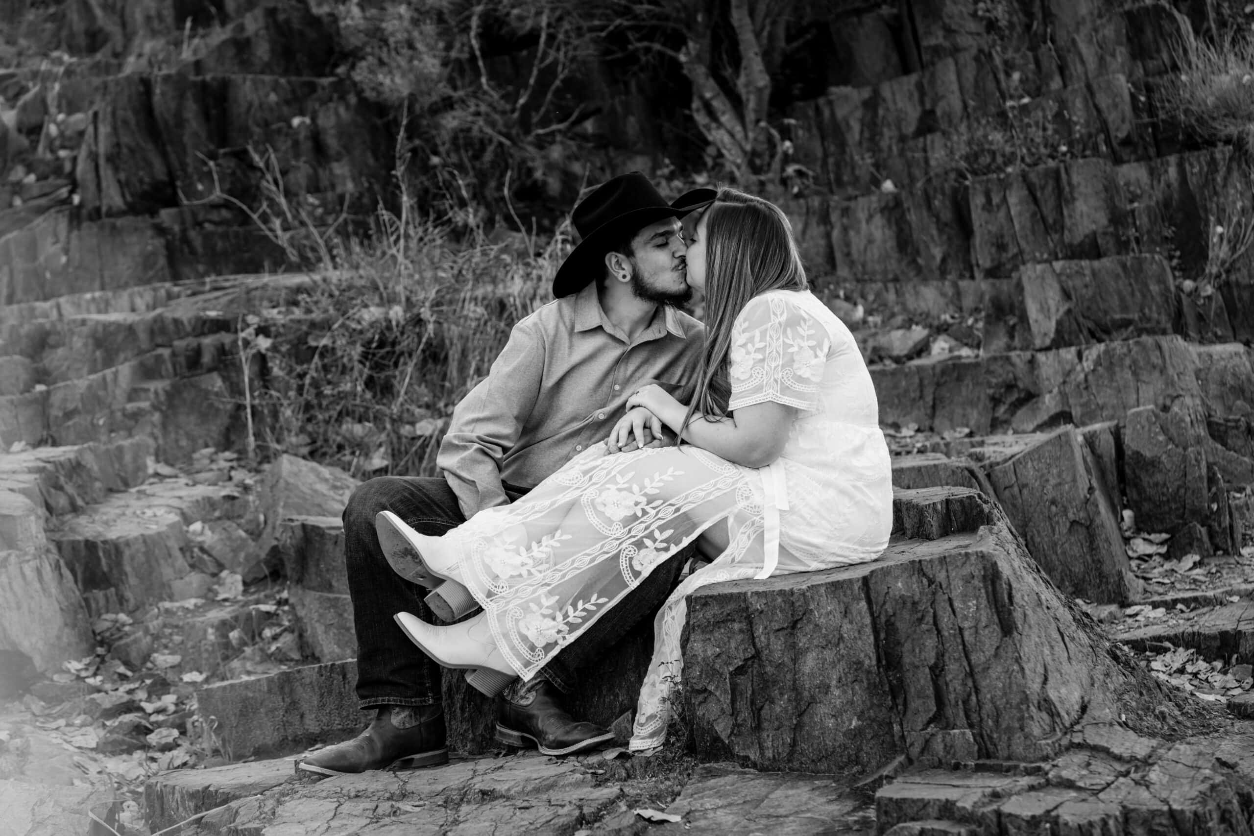 Couple kissing while sitting on rock formations, captured in a rustic and candid black and white style.