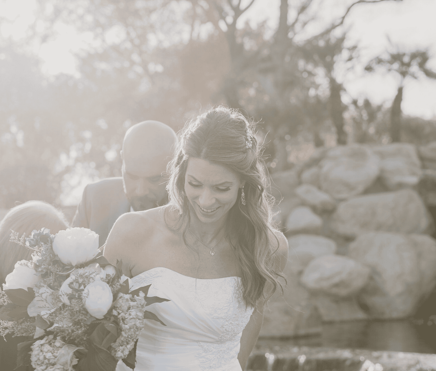 Bride smiling as she walks toward the ceremony holding bouquet, backlit by sunlight in a Virginia Beach garden setting
