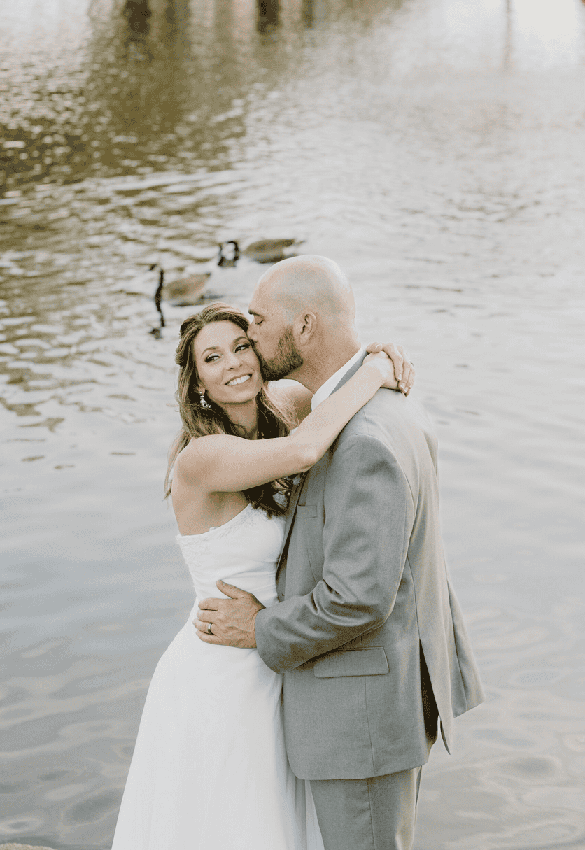 Bride smiling as groom kisses her by the lake with geese in the background, wedding photography in Virginia Beach
