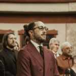 Stylish groom in maroon suit and glasses emotionally watching ceremony at outdoor wedding.