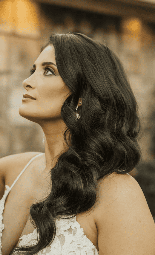 Elegant bride with wavy dark hair and sparkling earrings gazing upward in outdoor wedding portrait.