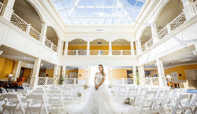 A bride posing beneath a glass-ceiling atrium at The Founders Inn, with ceremony chairs set up for a beautiful indoor wedding, captured by a Virginia Beach wedding photographer.