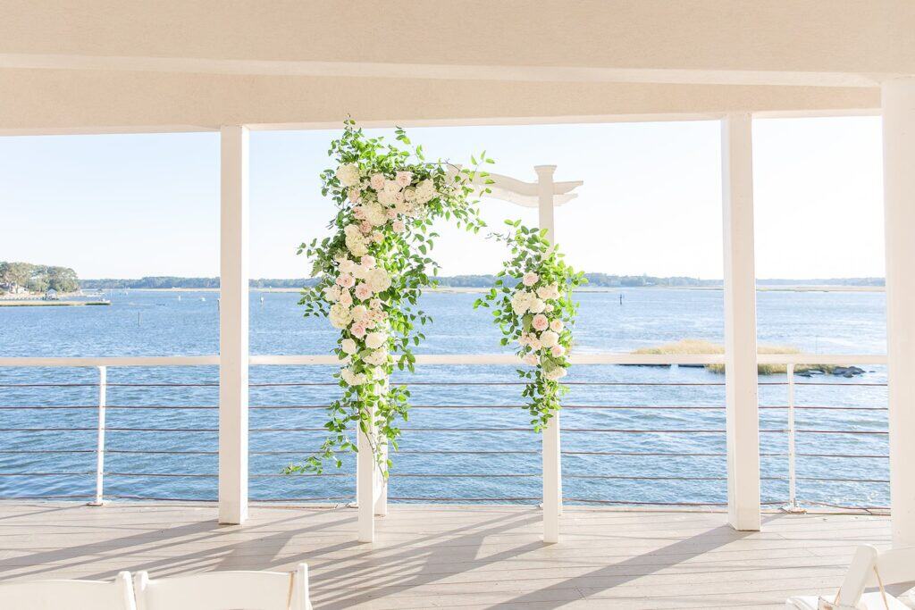 A scenic waterfront ceremony setup on The Lesner Inn’s deck overlooking the bay, with a floral arch frame captured by a Virginia Beach wedding photographer.