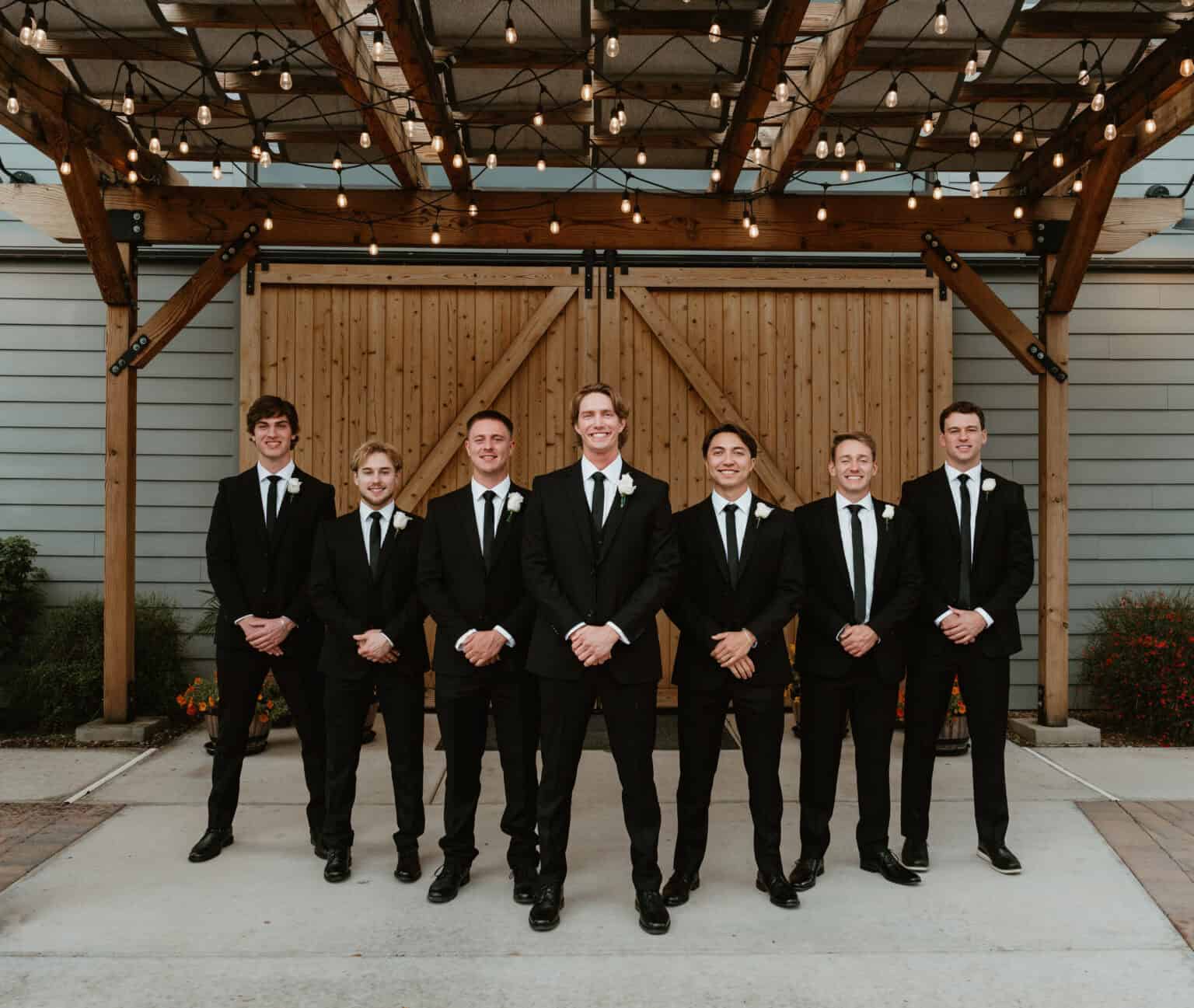Groom and seven groomsmen in black suits with white rose boutonnieres under string lights at a wedding, photographed by Captured in VA.
