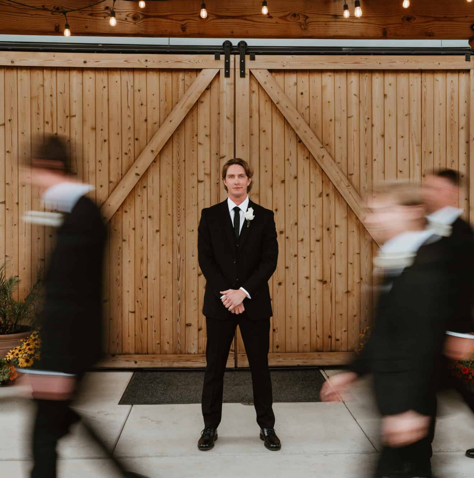 Groom standing still in a black suit as his groomsmen blur past in front of wooden barn doors, a creative wedding portrait by Captured in VA.