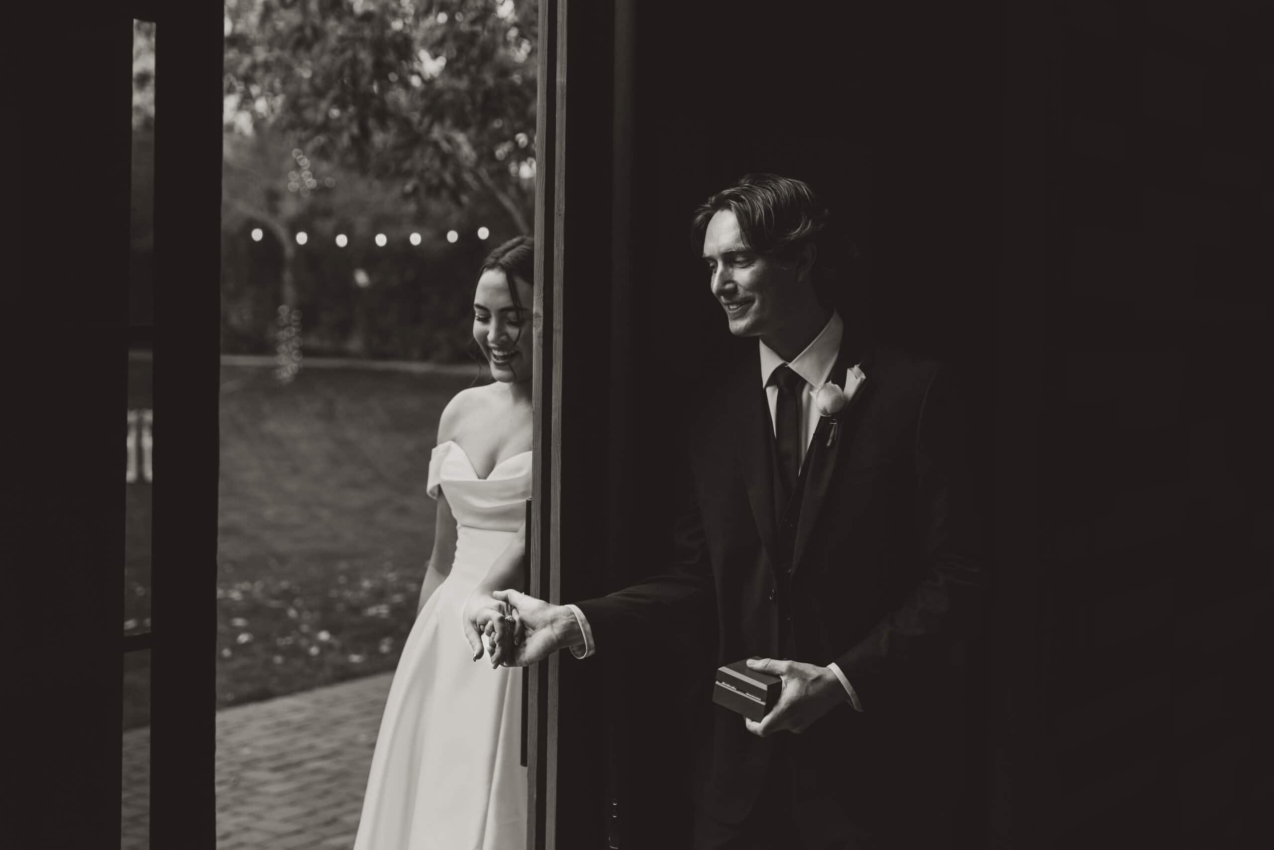 Black and white photo of a bride and groom sharing a private first touch around a corner before the ceremony, the groom holding a ring box, by Captured in VA.
