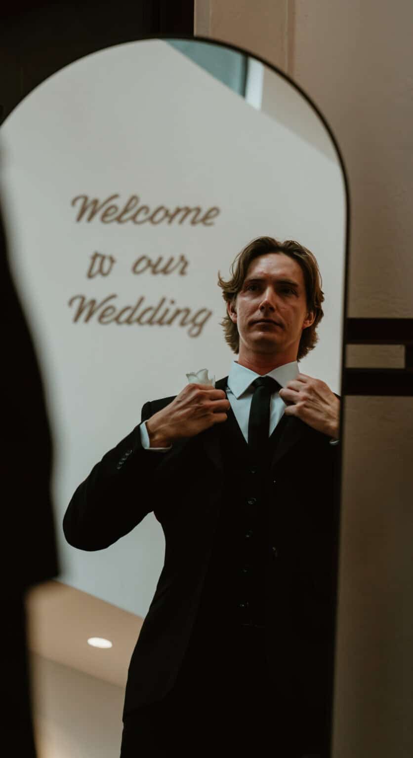Groom adjusting his tie in an arched mirror reflecting a 'Welcome to our Wedding' sign while getting ready, by Captured in VA.