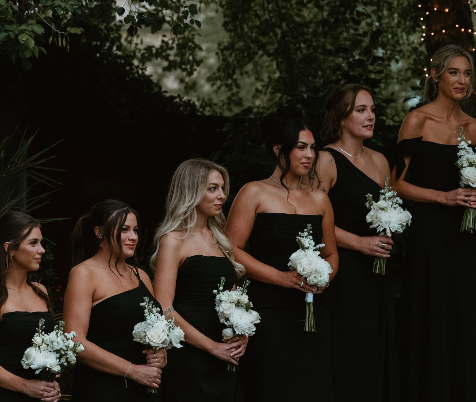 Bridesmaids in black gowns holding white rose bouquets in front of lush greenery at a wedding, photographed by Captured in VA.