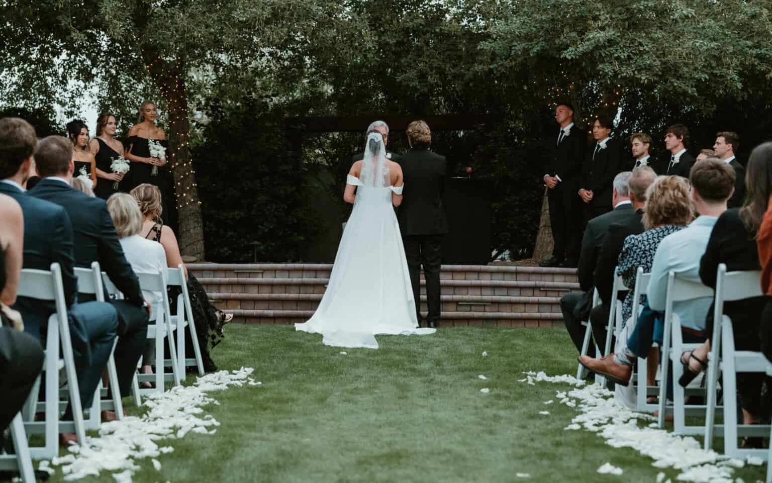 Bride in a lace veil facing her bridesmaids in black gowns during an outdoor garden wedding ceremony, by Captured in VA.