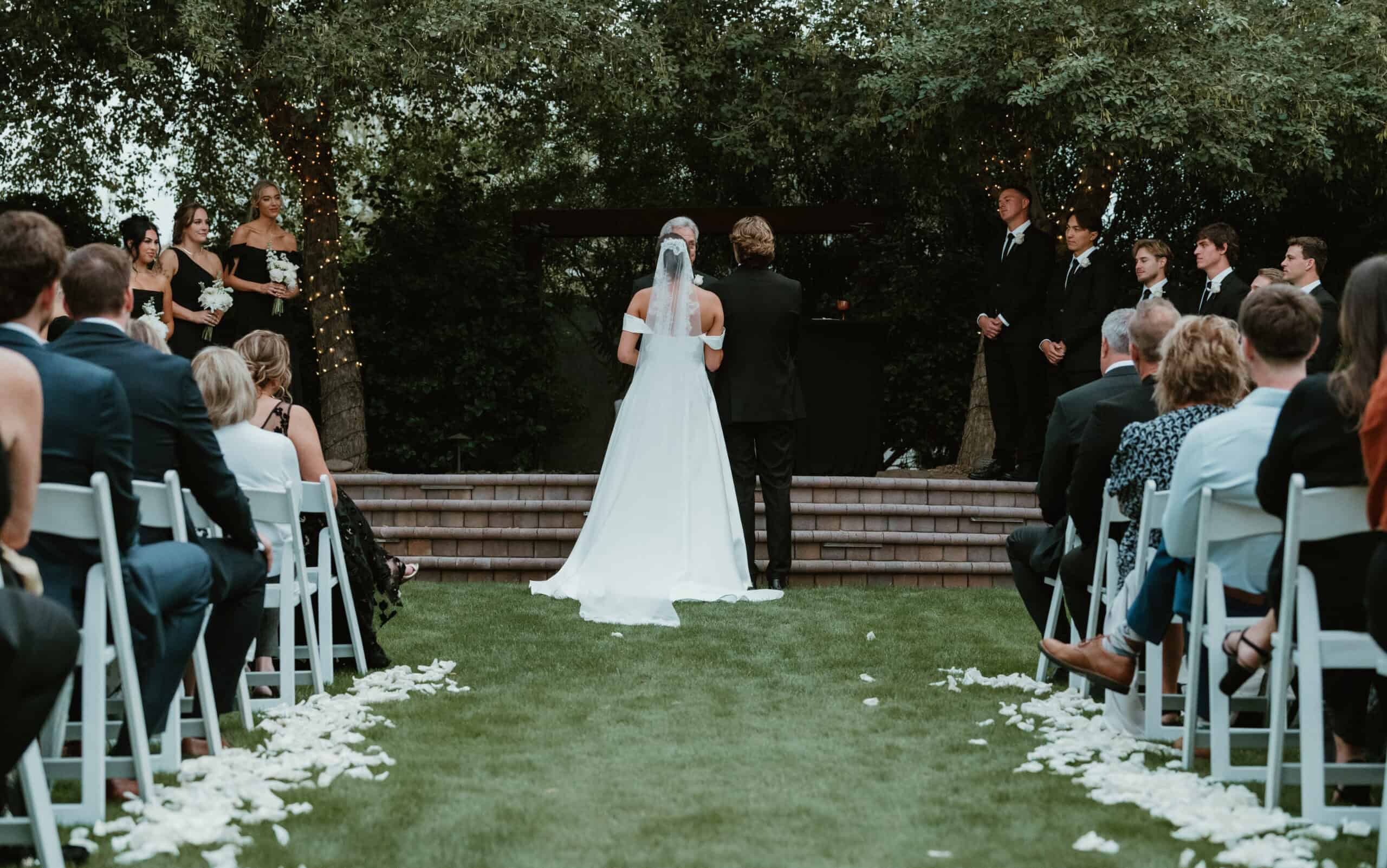 Bride in a lace veil facing her bridesmaids in black gowns during an outdoor garden wedding ceremony, by Captured in VA.