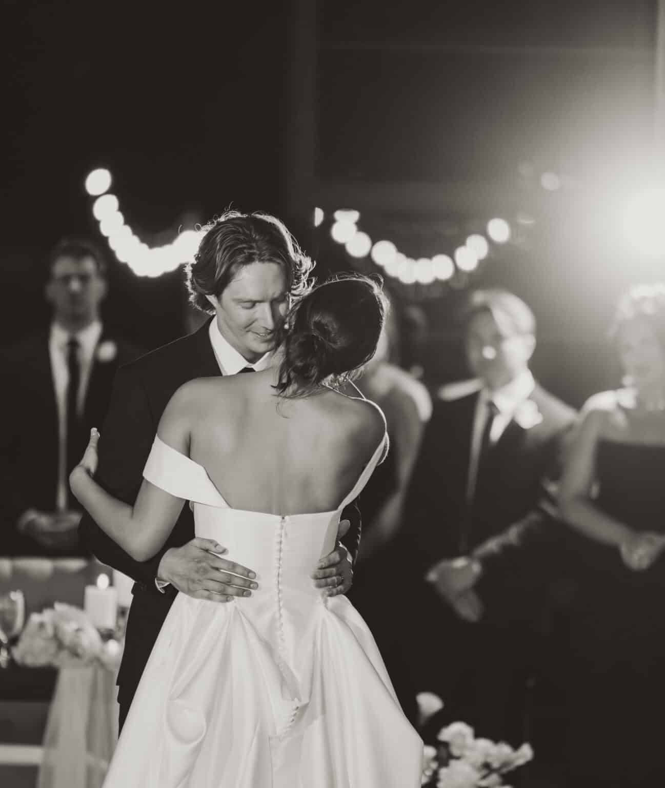 Black and white photo of the bride and groom sharing their first dance under string lights as guests watch, by Captured in VA.