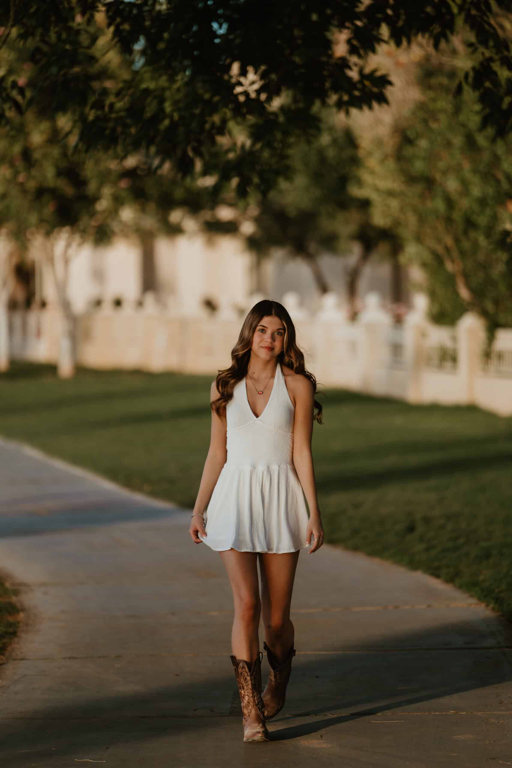 High school senior walking toward the camera in a white dress and cowboy boots along a tree-lined street at golden hour, by Captured in VA.