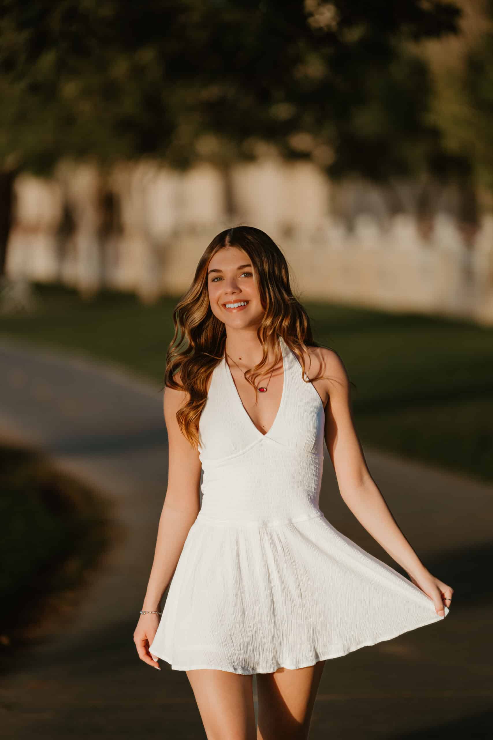 Smiling high school senior holding out the skirt of her white halter dress in warm golden-hour light, a senior portrait by Captured in VA.