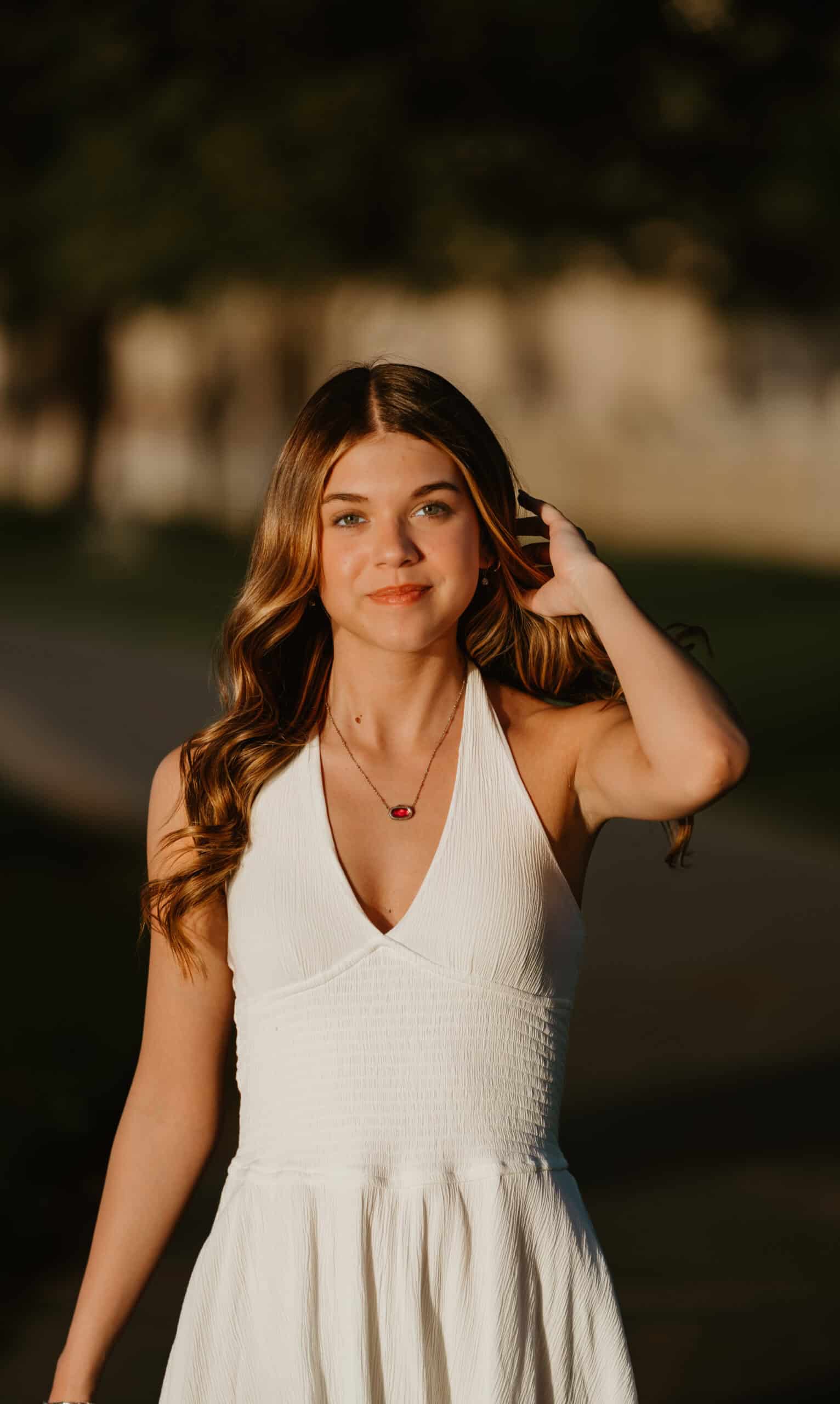 Close-up senior portrait of a girl with her hand in her hair, wearing a white halter dress with a red pendant necklace at golden hour, by Captured in VA.