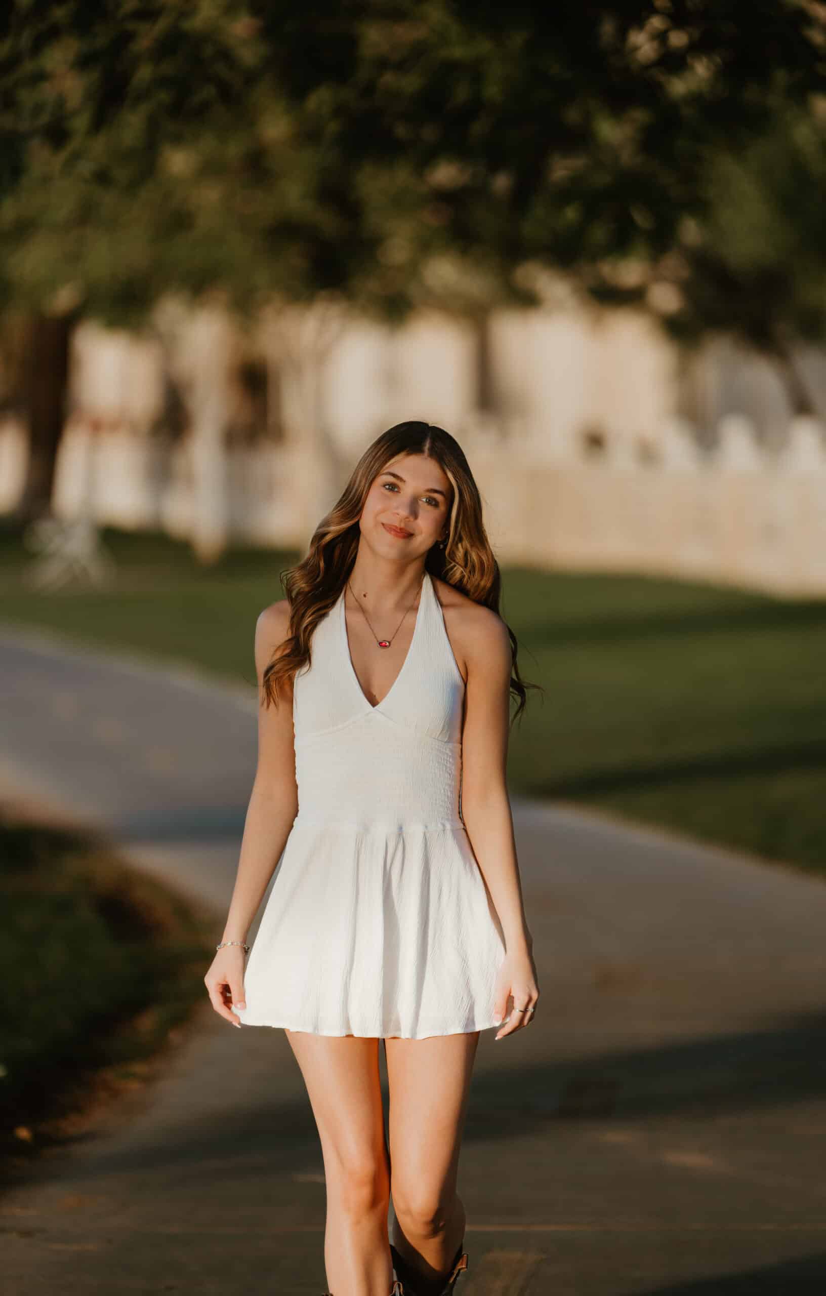 High school senior in a white halter dress walking down a sunlit, tree-lined path at golden hour, by Captured in VA.