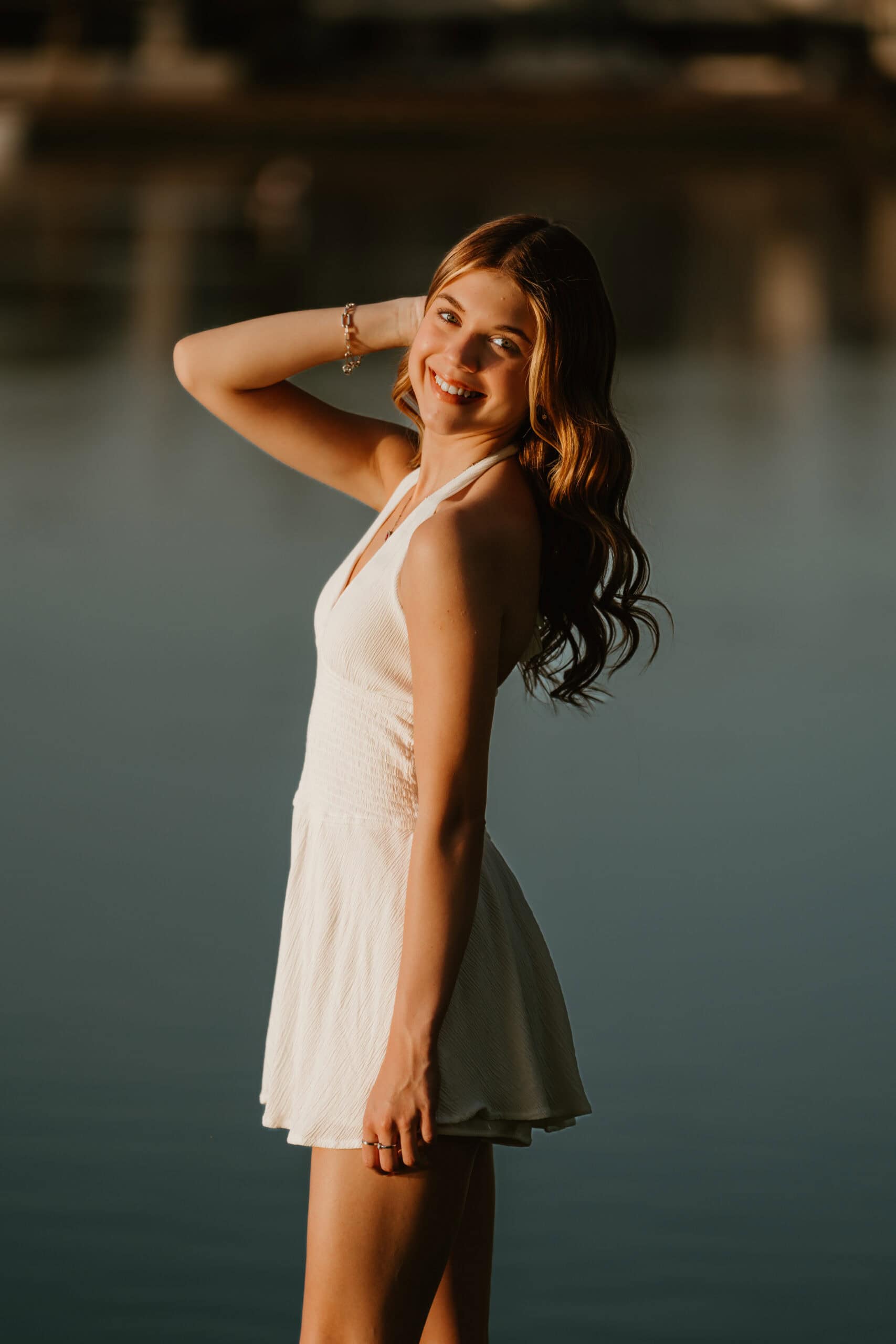 High school senior smiling over her shoulder in a white dress beside the water at golden hour, a senior portrait by Captured in VA.