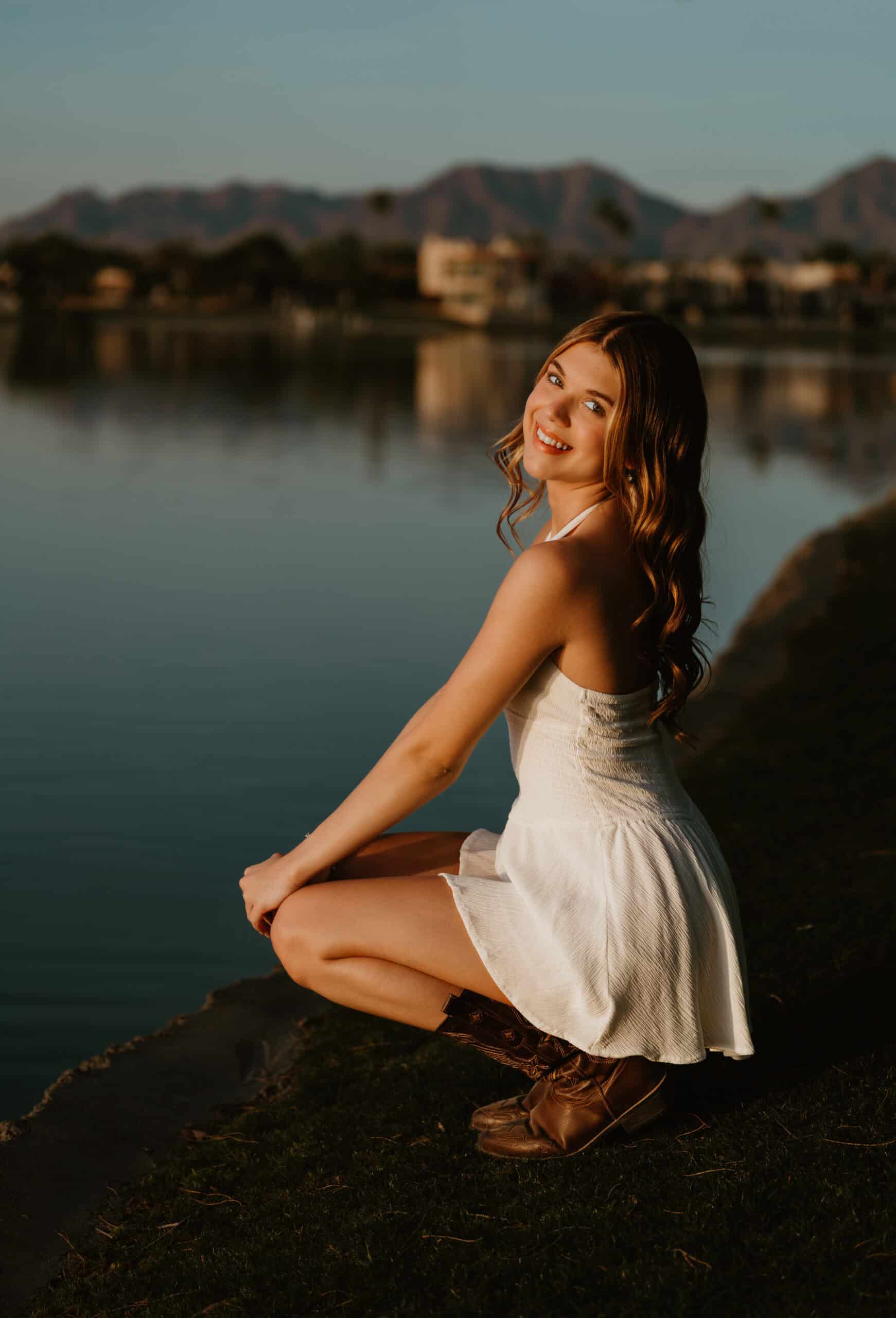 High school senior in a white dress and cowboy boots crouching beside a calm lake at sunset with mountains behind, by Captured in VA.