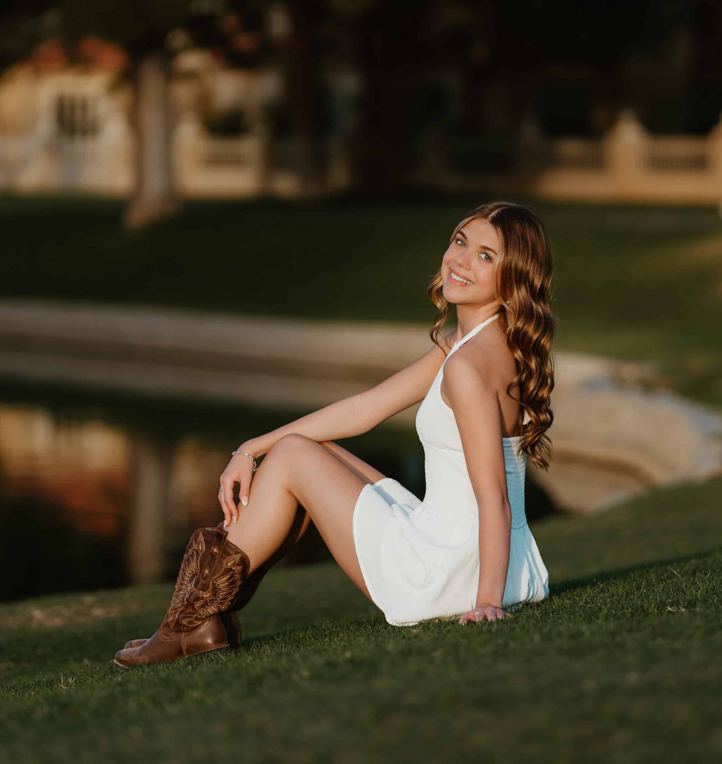 High school senior in a white dress and cowboy boots sitting on the grass beside a pond at golden hour, smiling over her shoulder, by Captured in VA.
