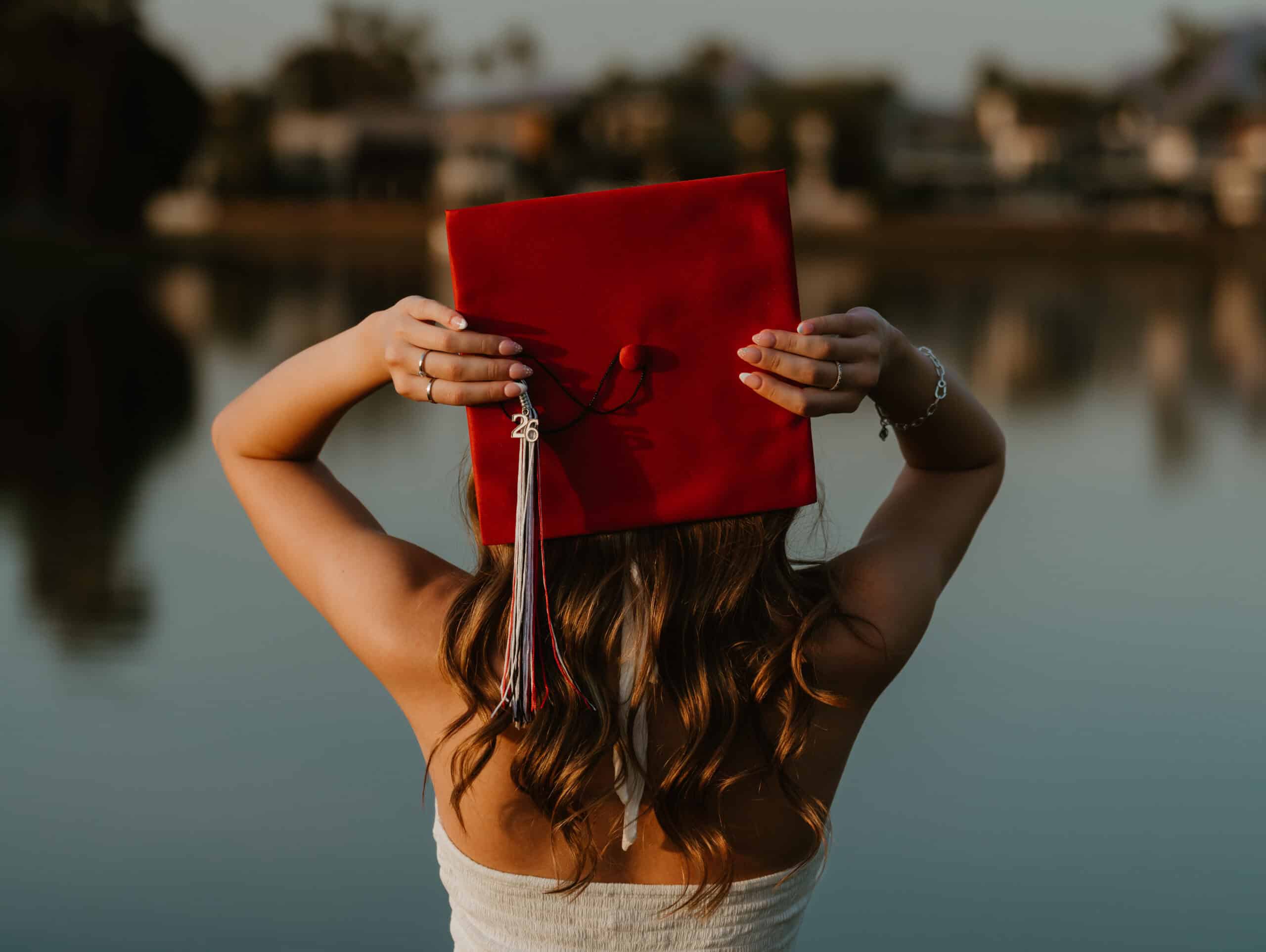Back view of a Class of 2026 senior raising a red graduation cap with a red and white tassel in front of a calm lake, by Captured in VA.
