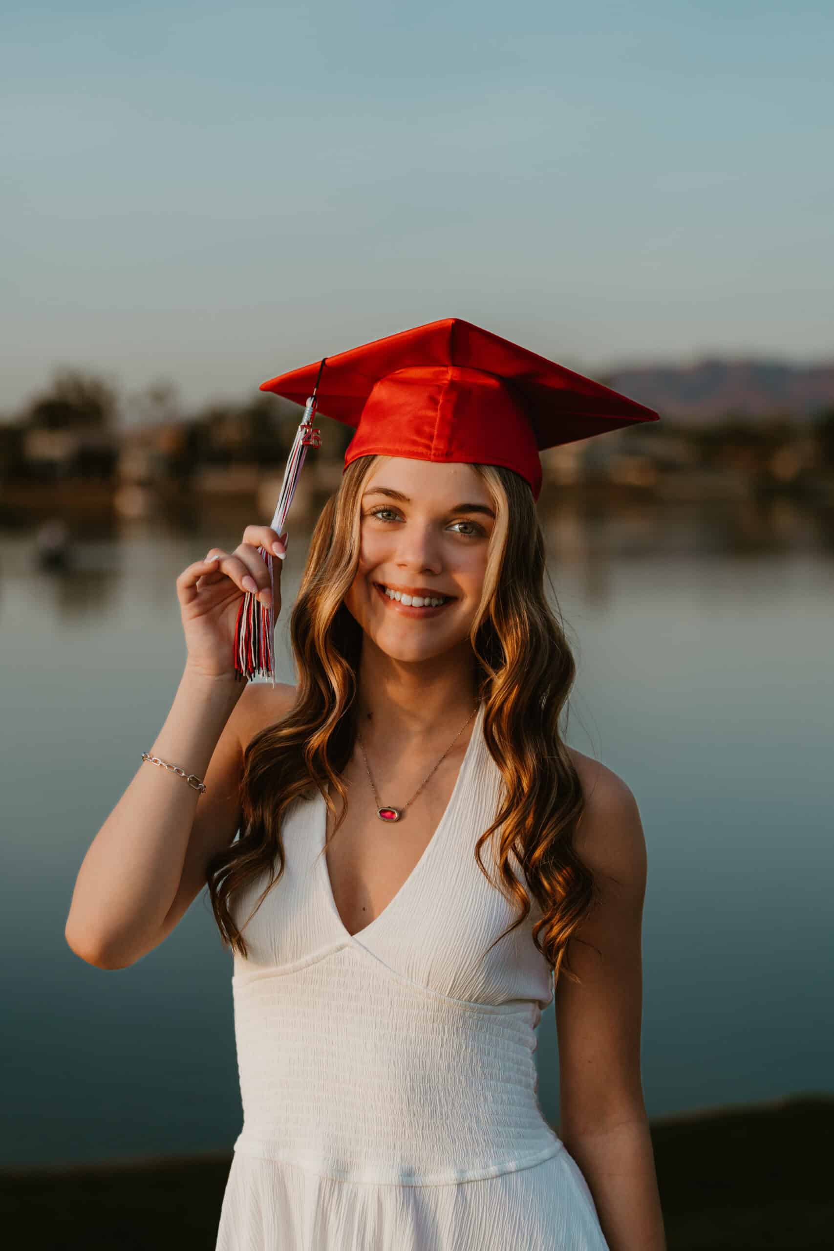Smiling high school senior wearing a red graduation cap and holding her '26' tassel beside a lake at sunset, by Captured in VA.
