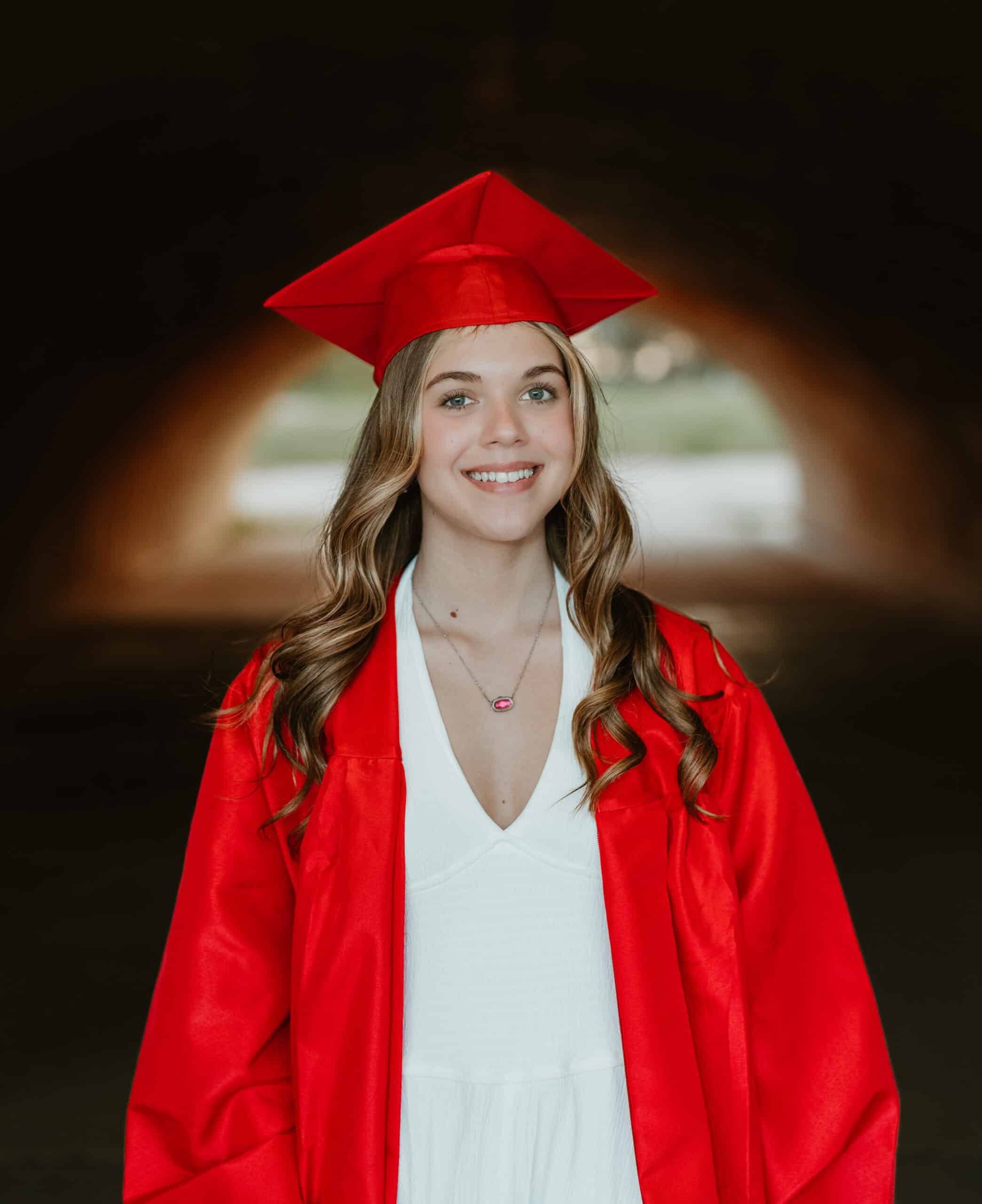 Class of 2026 senior in a red cap and gown over a white dress, framed by a soft-lit archway, by Captured in VA.