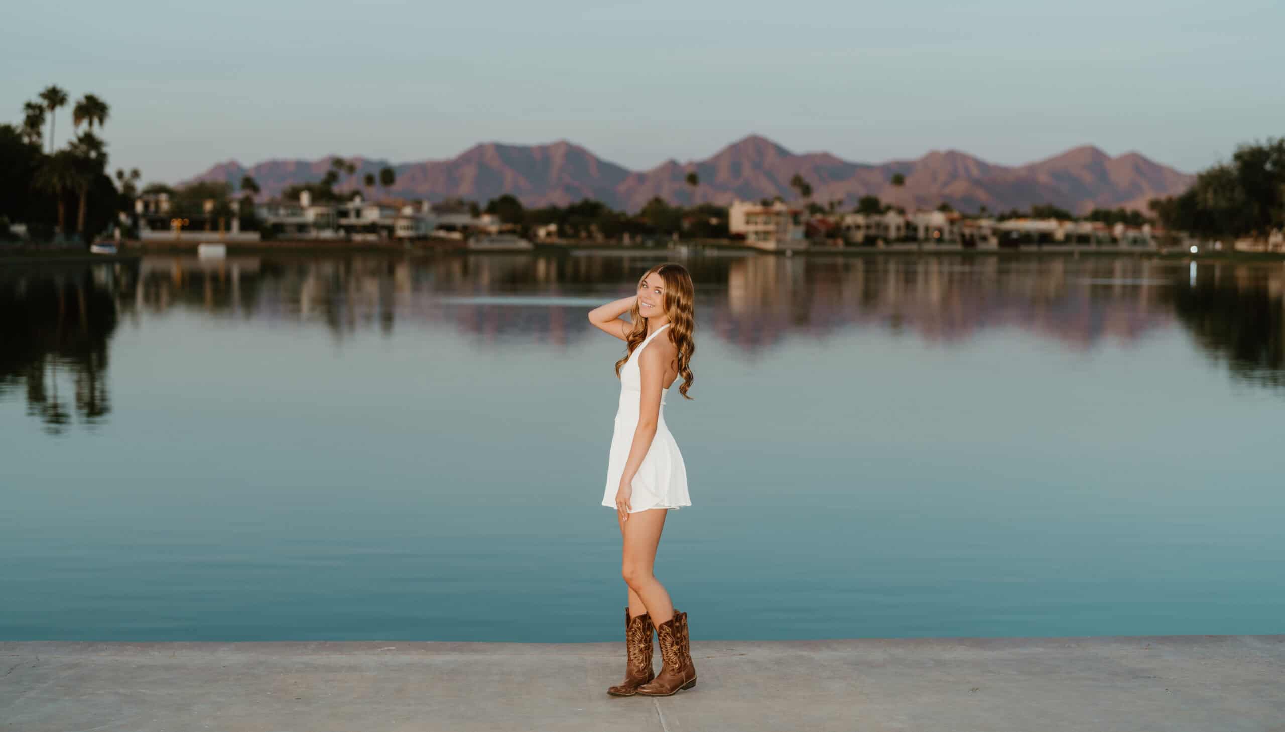 High school senior in a white dress and cowboy boots standing by a lake with desert mountains and palm trees behind, by Captured in VA.