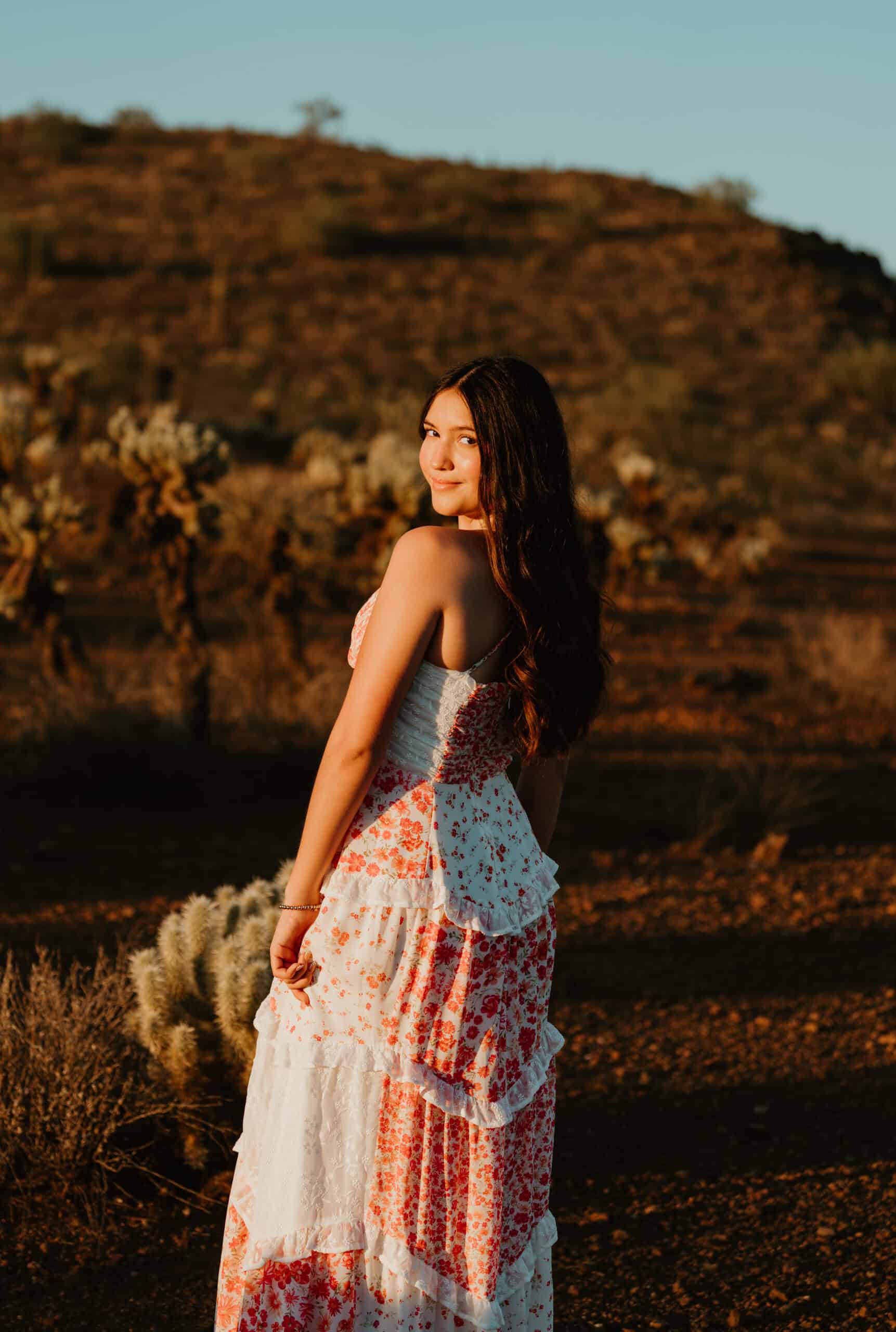 High school senior in a floral maxi dress glancing back amid cholla cactus and desert hills at golden hour, by Captured in VA.
