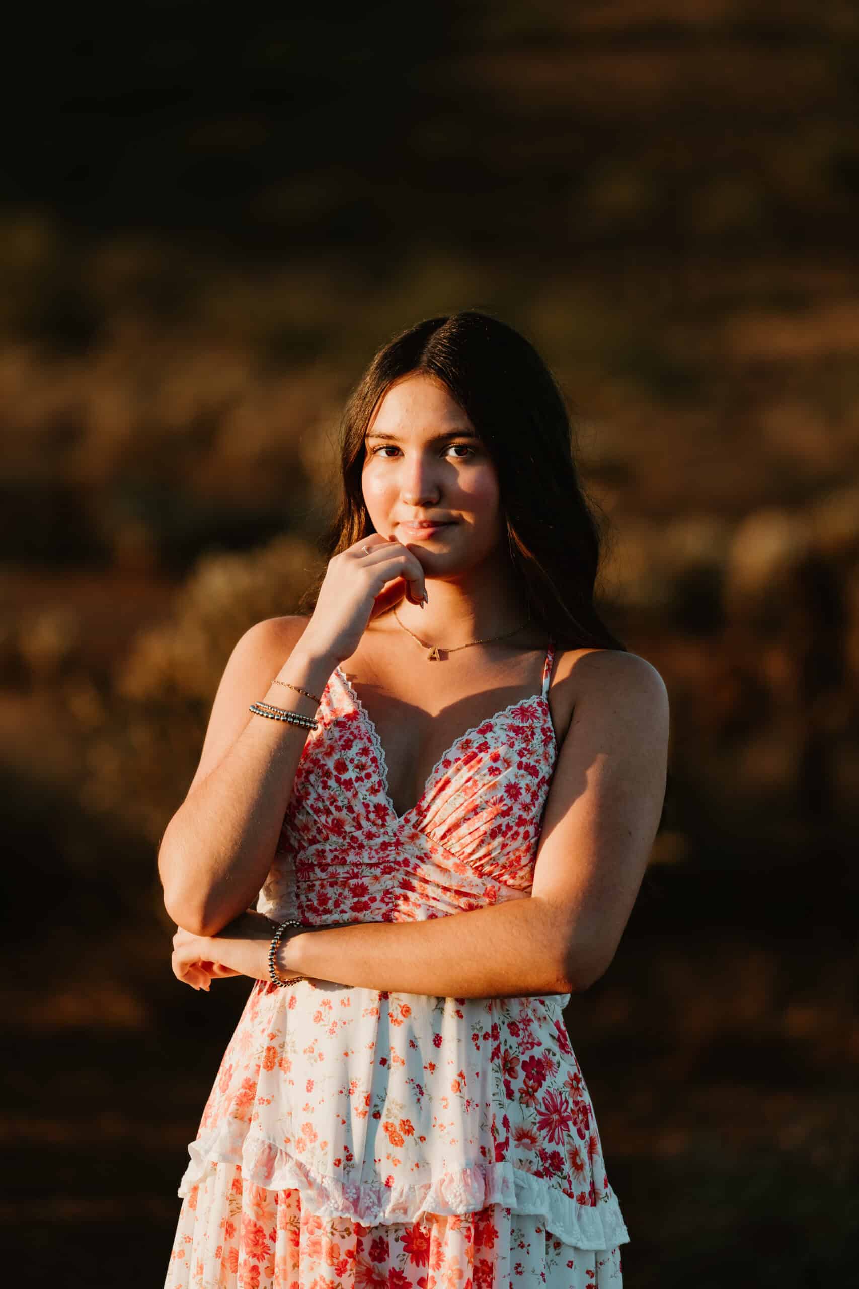 Close-up senior portrait of a girl in a red floral dress resting her hand near her chin in warm sunset light, by Captured in VA.