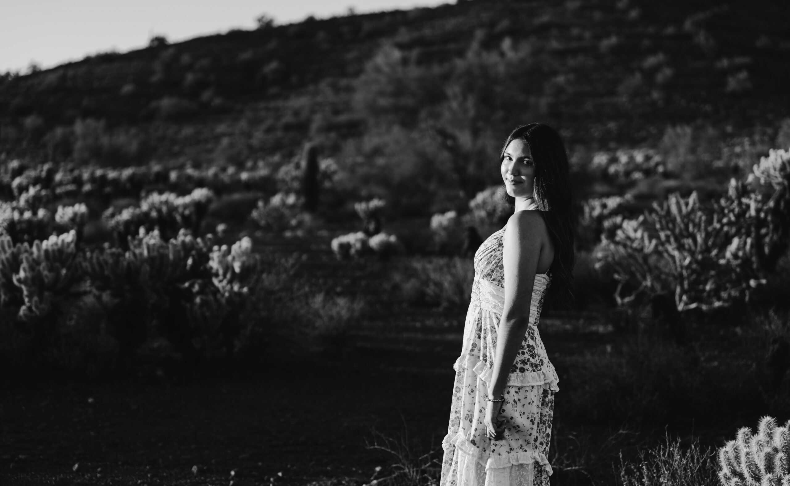 Black and white senior portrait of a girl in a floral maxi dress standing in profile among desert brush and cactus, by Captured in VA.