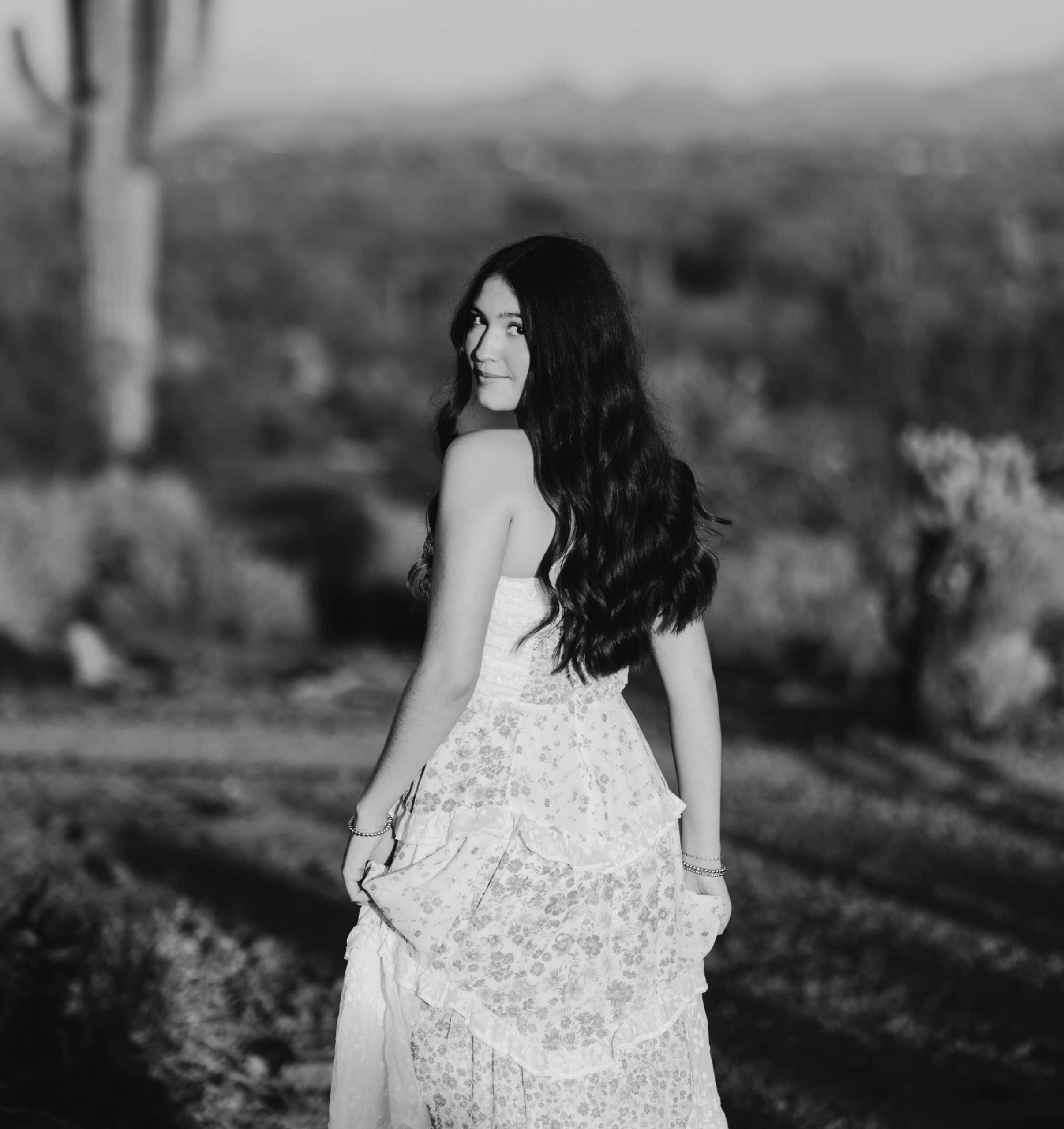 Black and white senior portrait of a girl in a floral maxi dress looking over her shoulder in the desert, by Captured in VA.
