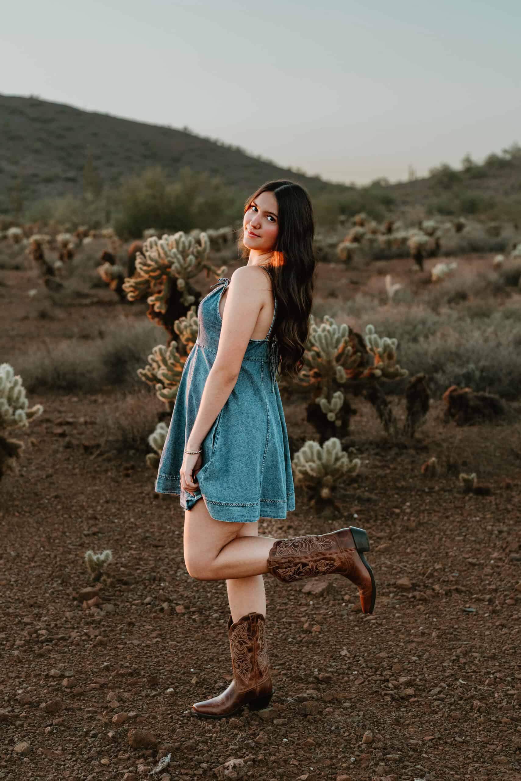 High school senior in a denim dress kicking up a cowboy boot in a cholla cactus desert with a hill behind at golden hour, by Captured in VA.