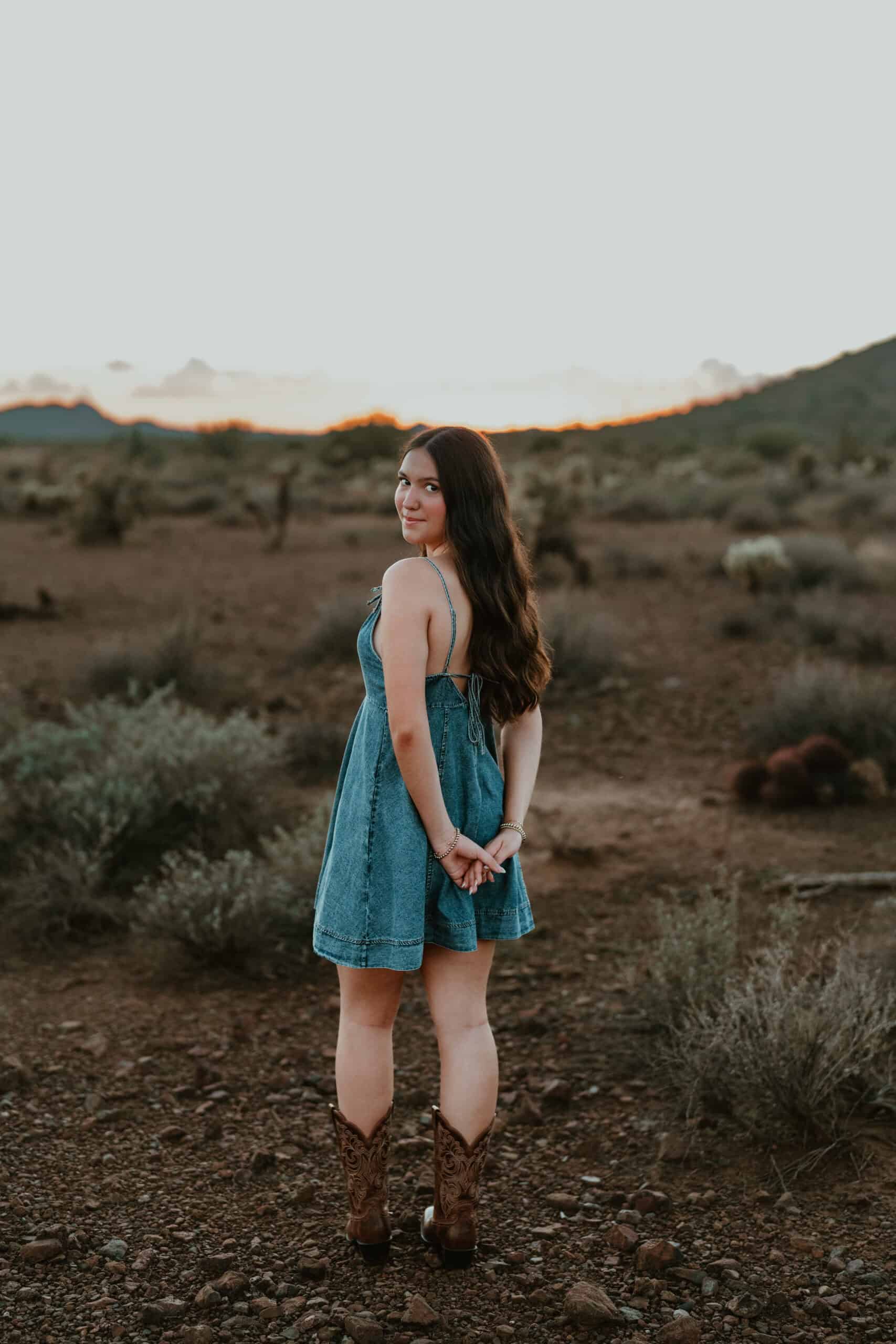 Playful high school senior in a denim dress kicking up one cowboy boot among cholla cactus at dusk, by Captured in VA.