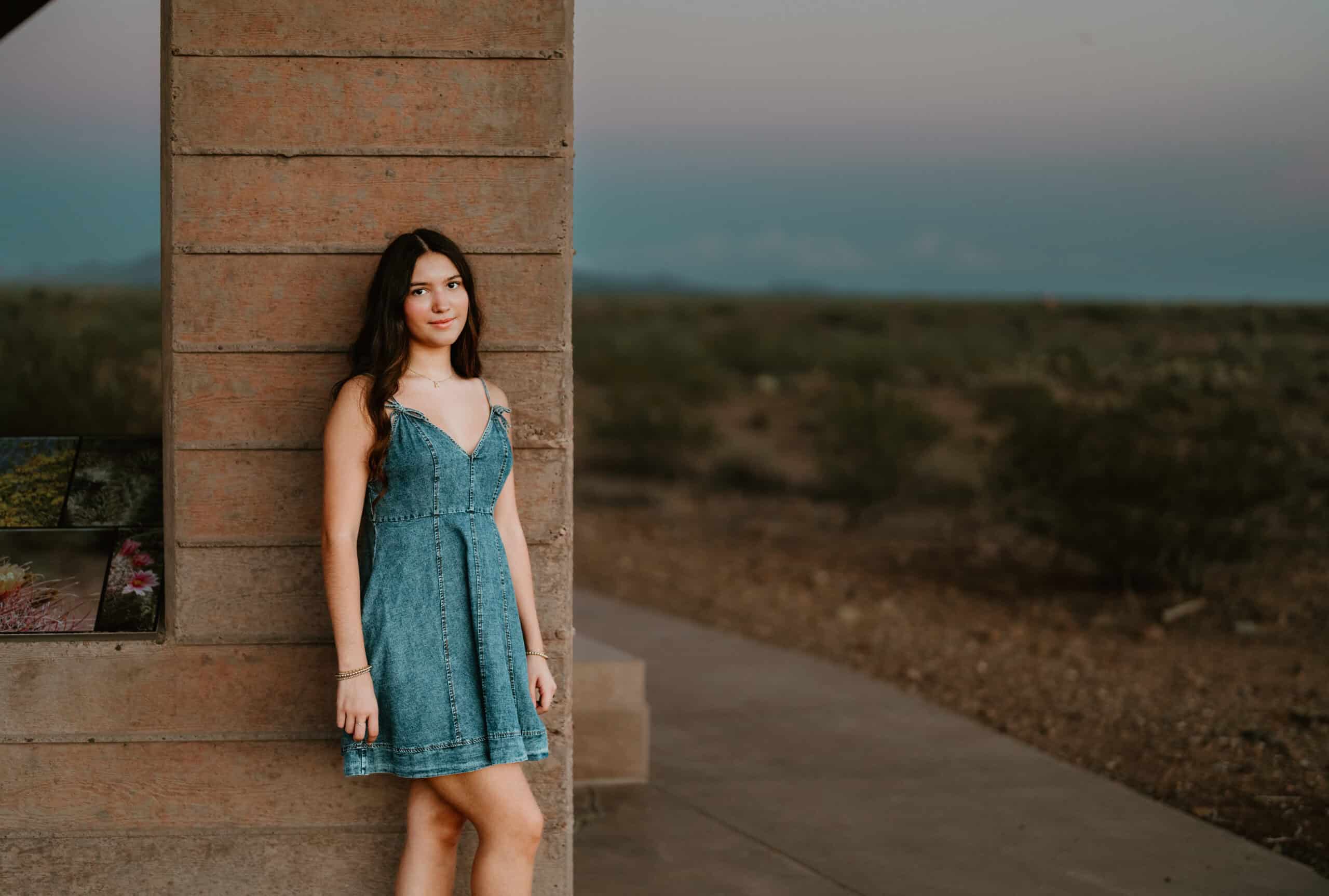 High school senior in a denim dress leaning against a wall at twilight with the desert behind her, by Captured in VA.