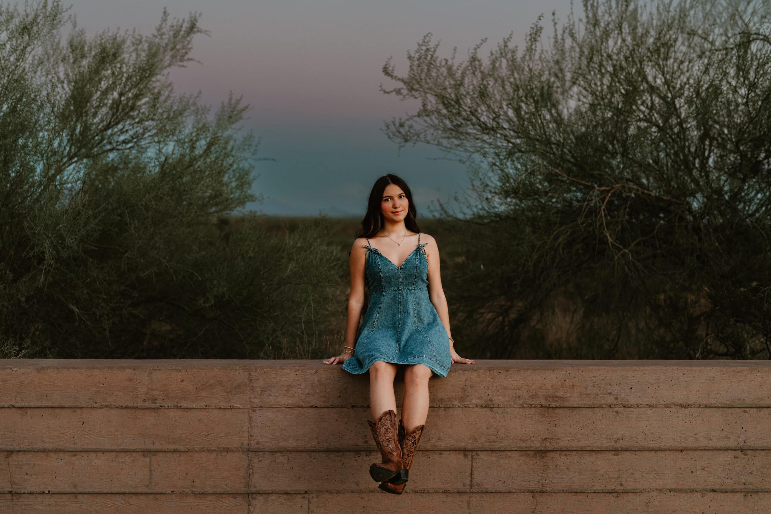 High school senior in a denim dress and cowboy boots sitting on a low wall with desert brush behind at dusk, by Captured in VA.