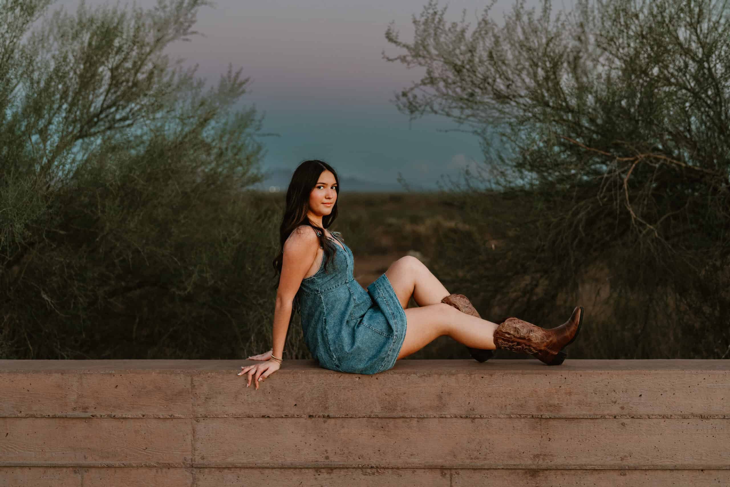 High school senior in a denim dress reclining along a low wall with her cowboy boots crossed, desert dusk behind, by Captured in VA.