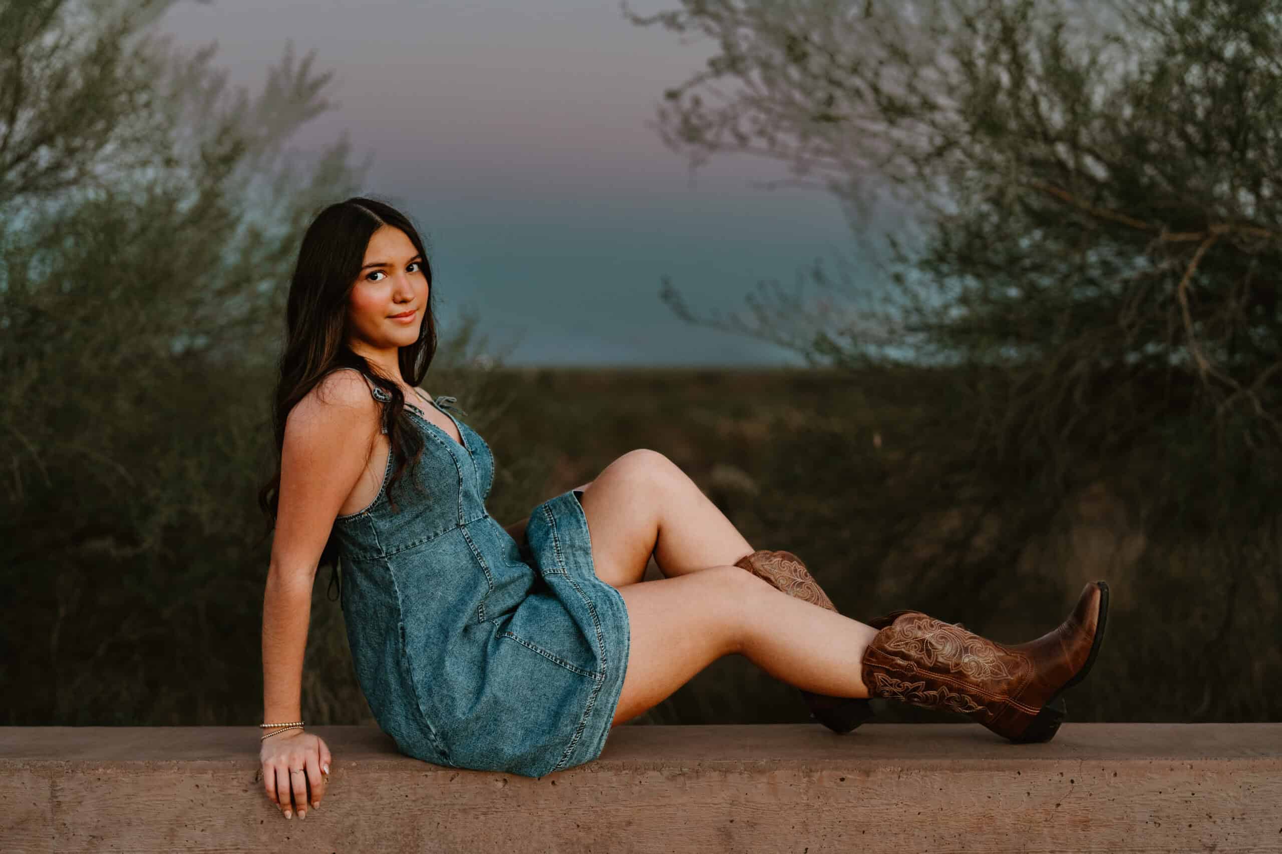 High school senior in a denim dress and embroidered cowboy boots seated on a wall at blue hour, looking at the camera, by Captured in VA.