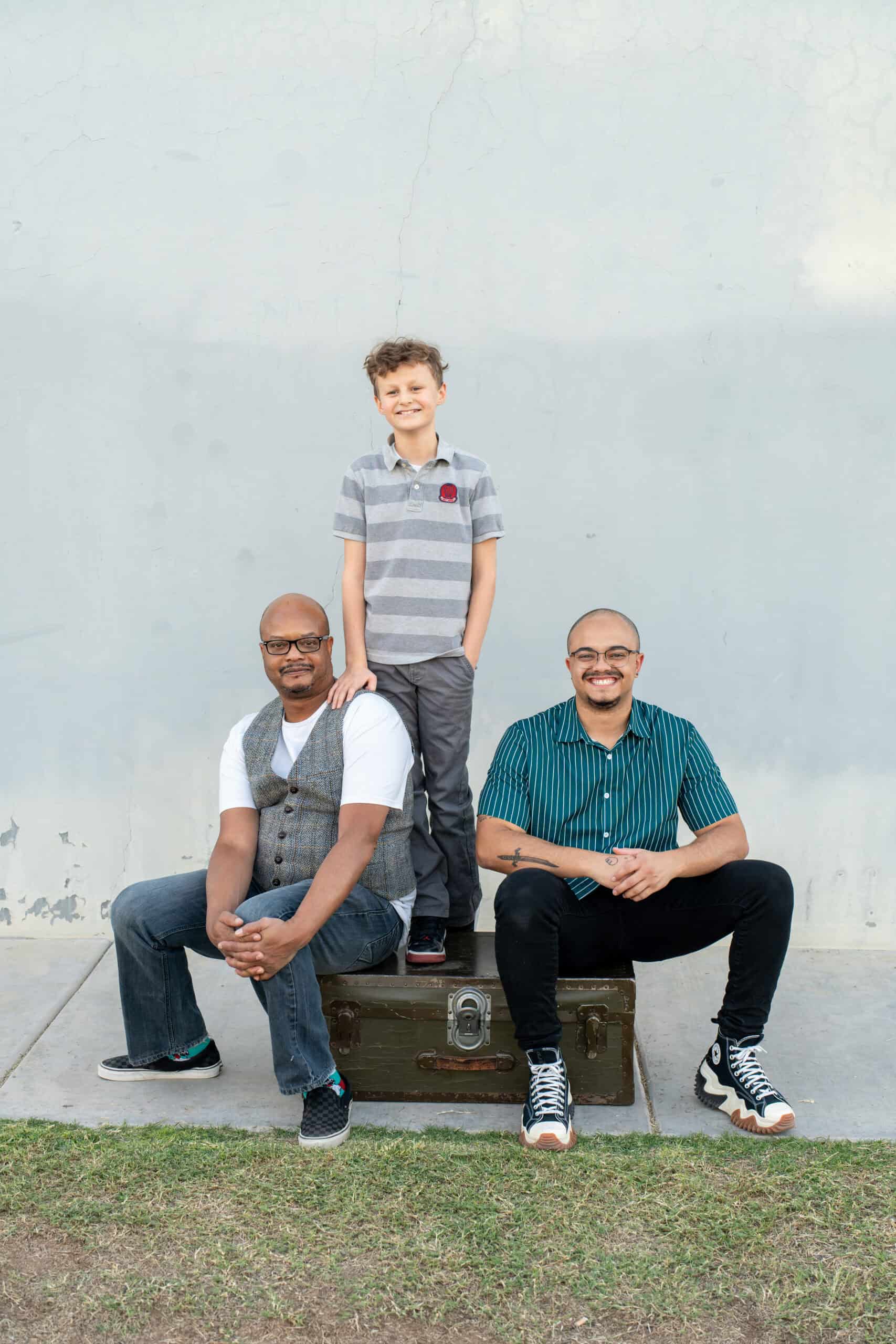 Two men and a young boy posing with a vintage trunk against a stucco wall during a family portrait session, by Captured in VA.
