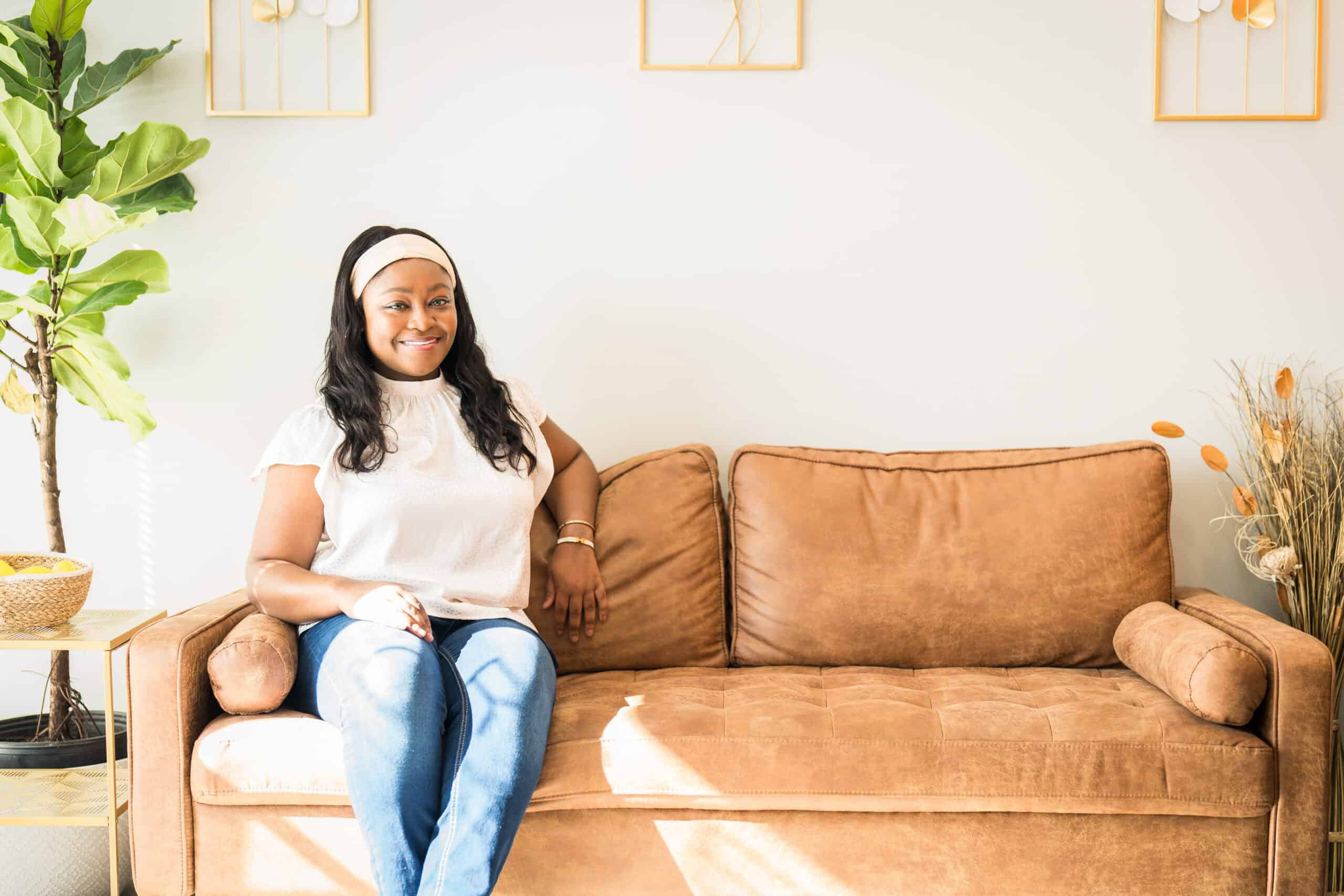 Woman in a white top and jeans seated on a tan leather couch beside a fiddle-leaf plant, a lifestyle portrait by Captured in VA.