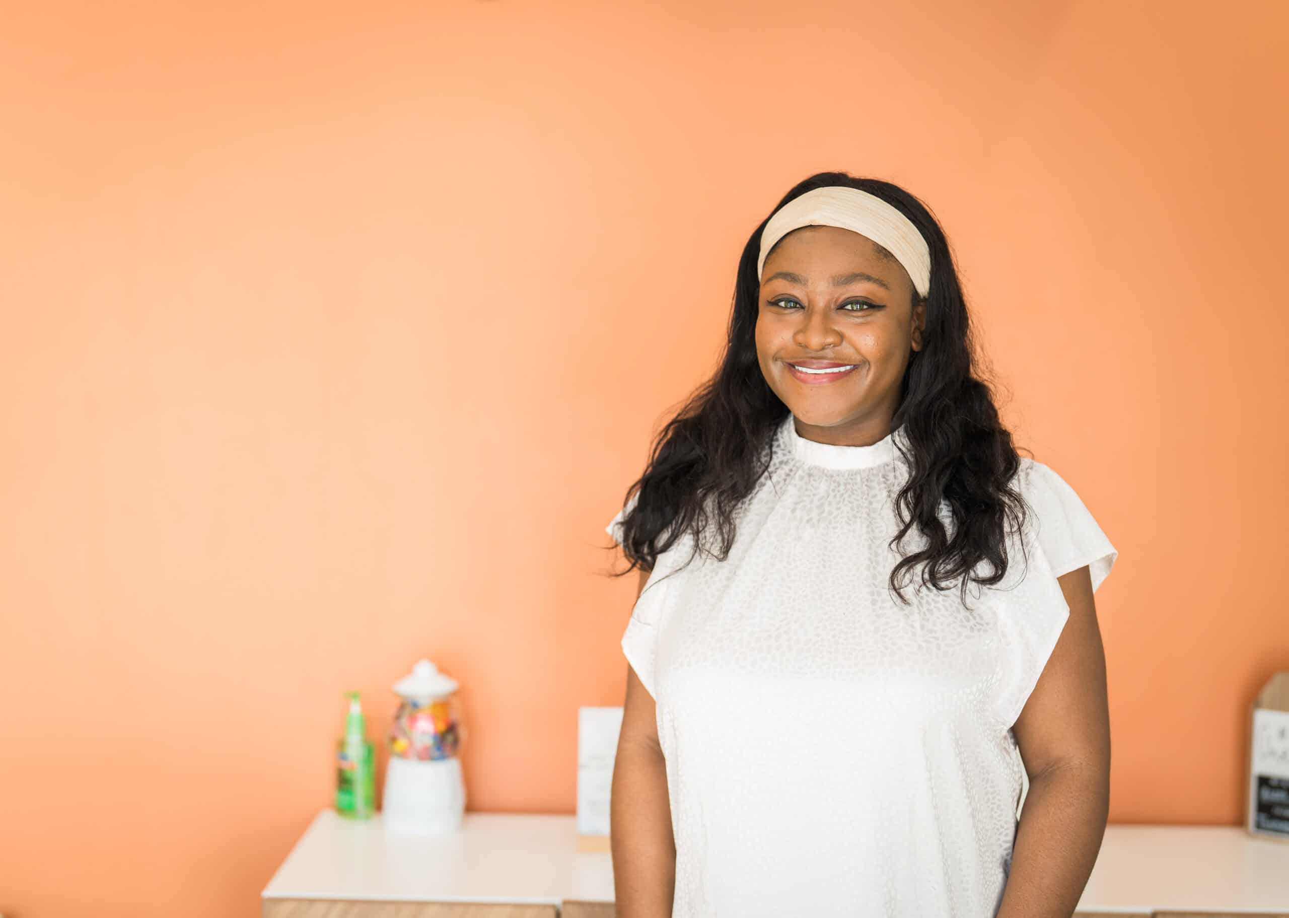 Smiling woman in a white top and headband standing against an orange wall in her business, a portrait by Captured in VA.
