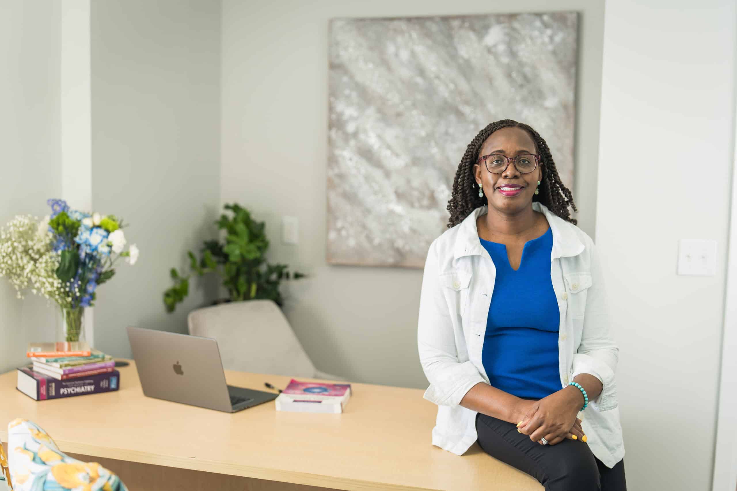Mental health clinician in a blue top and white jacket seated on a desk with a laptop and psychiatry books in her office, a branding portrait by Captured in VA.