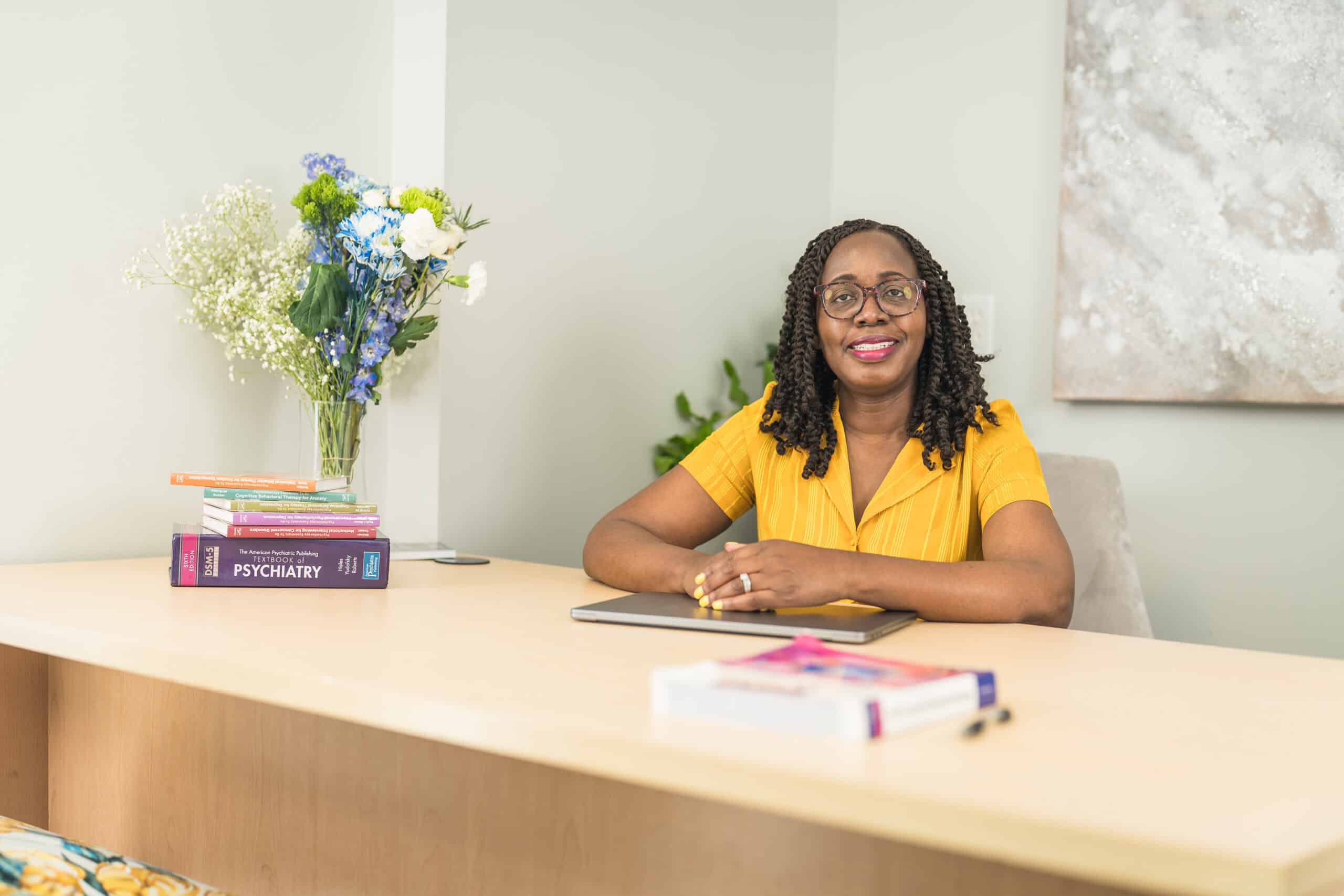 Psychiatry provider in a yellow top seated at a desk with a laptop and stacked psychiatry textbooks, a branding portrait by Captured in VA.